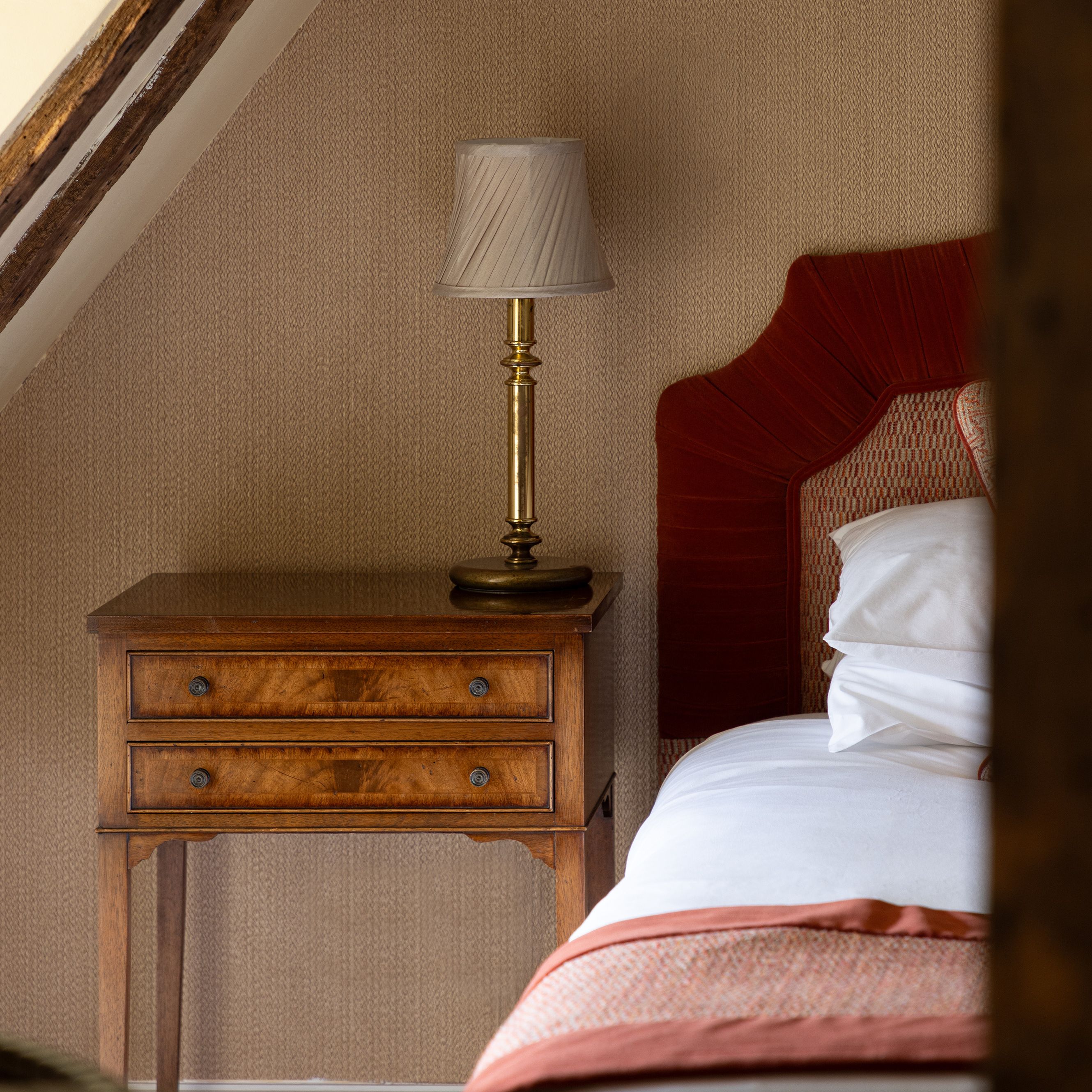 A cozy bedroom corner with a wooden nightstand, brass lamp, and part of a bed with a red headboard.