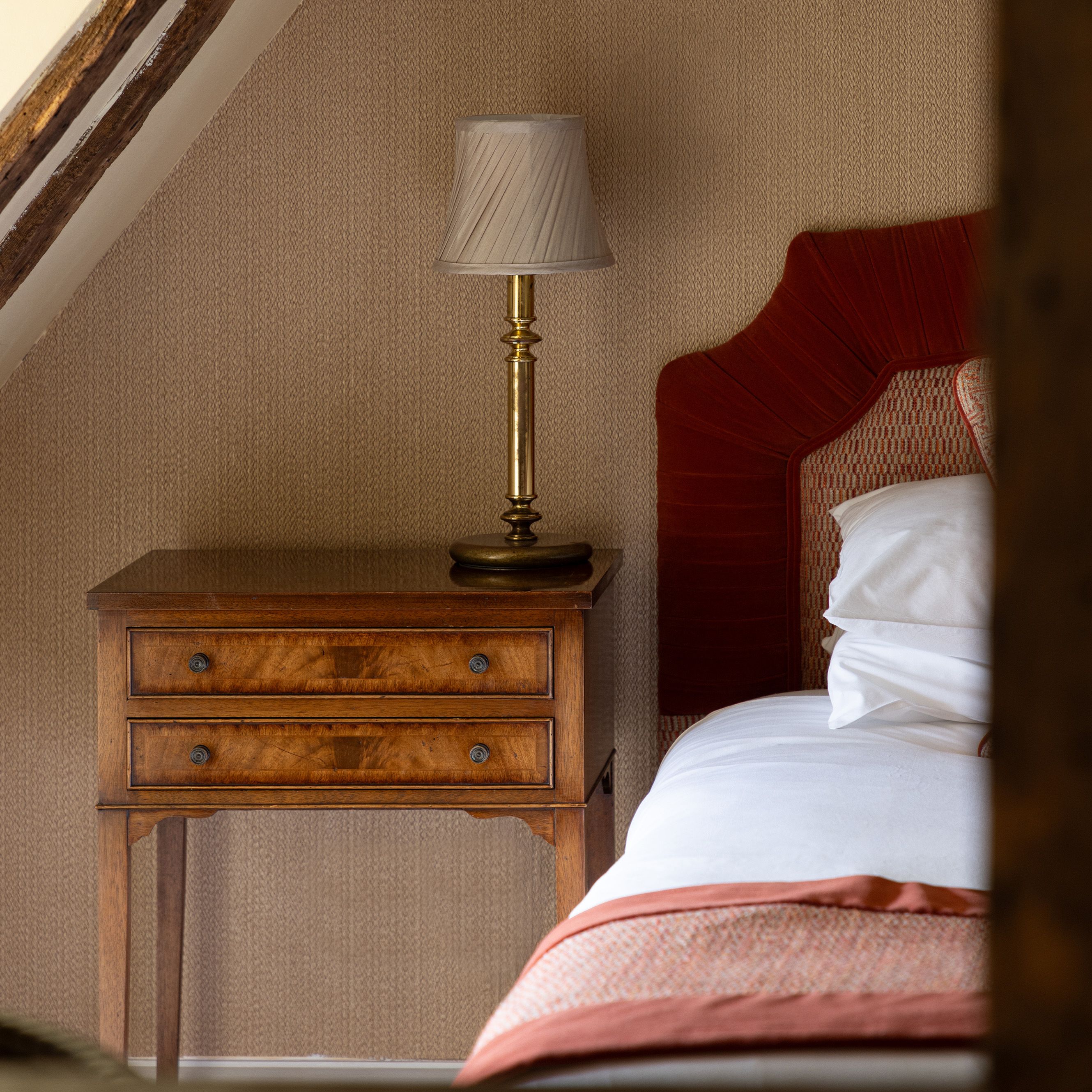 A cozy bedroom corner with a wooden nightstand, brass lamp, and part of a bed with a red headboard.