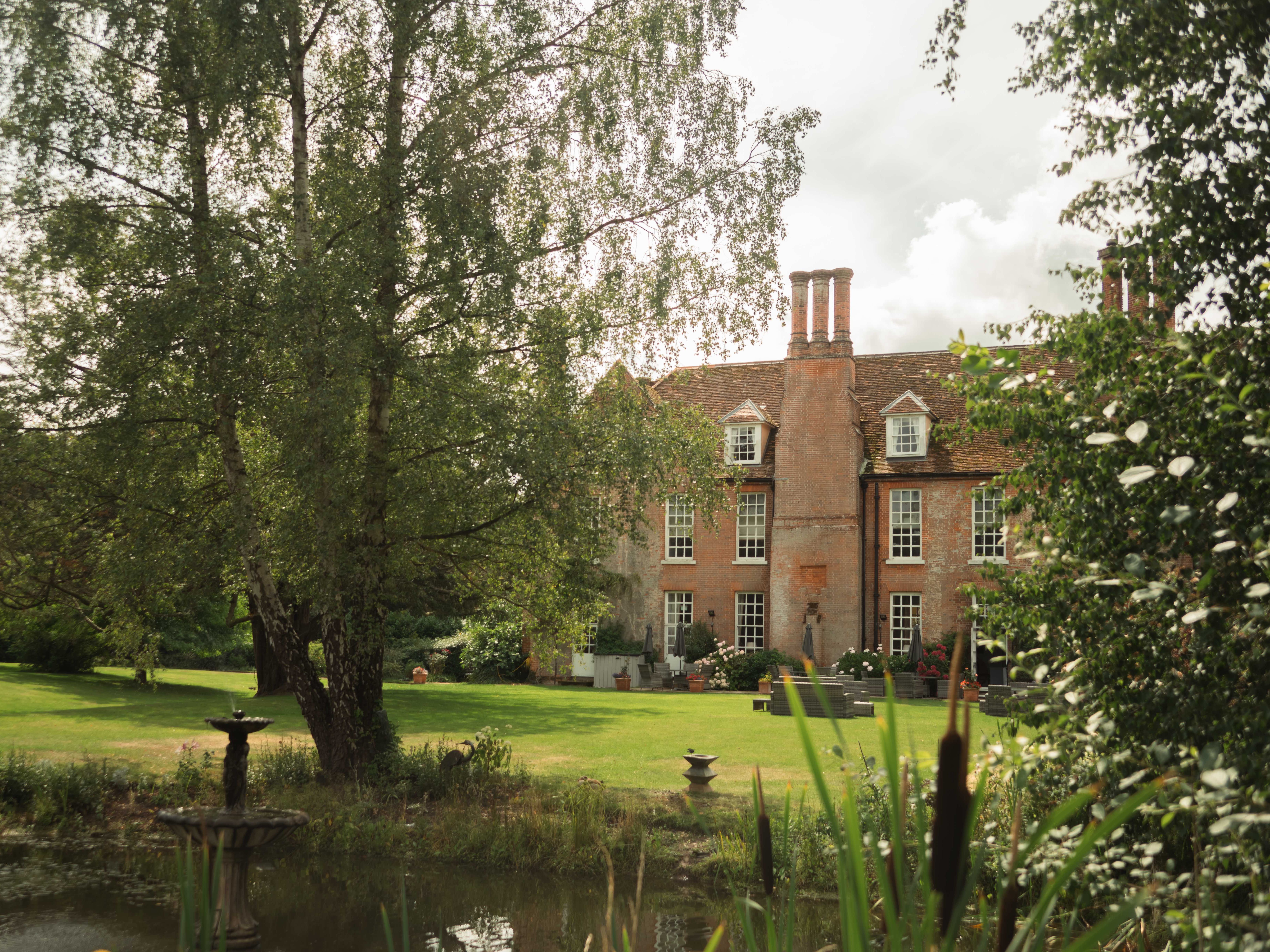 Large brick manor house with multiple chimneys, surrounded by lush green gardens and a pond with a fountain in the foreground.