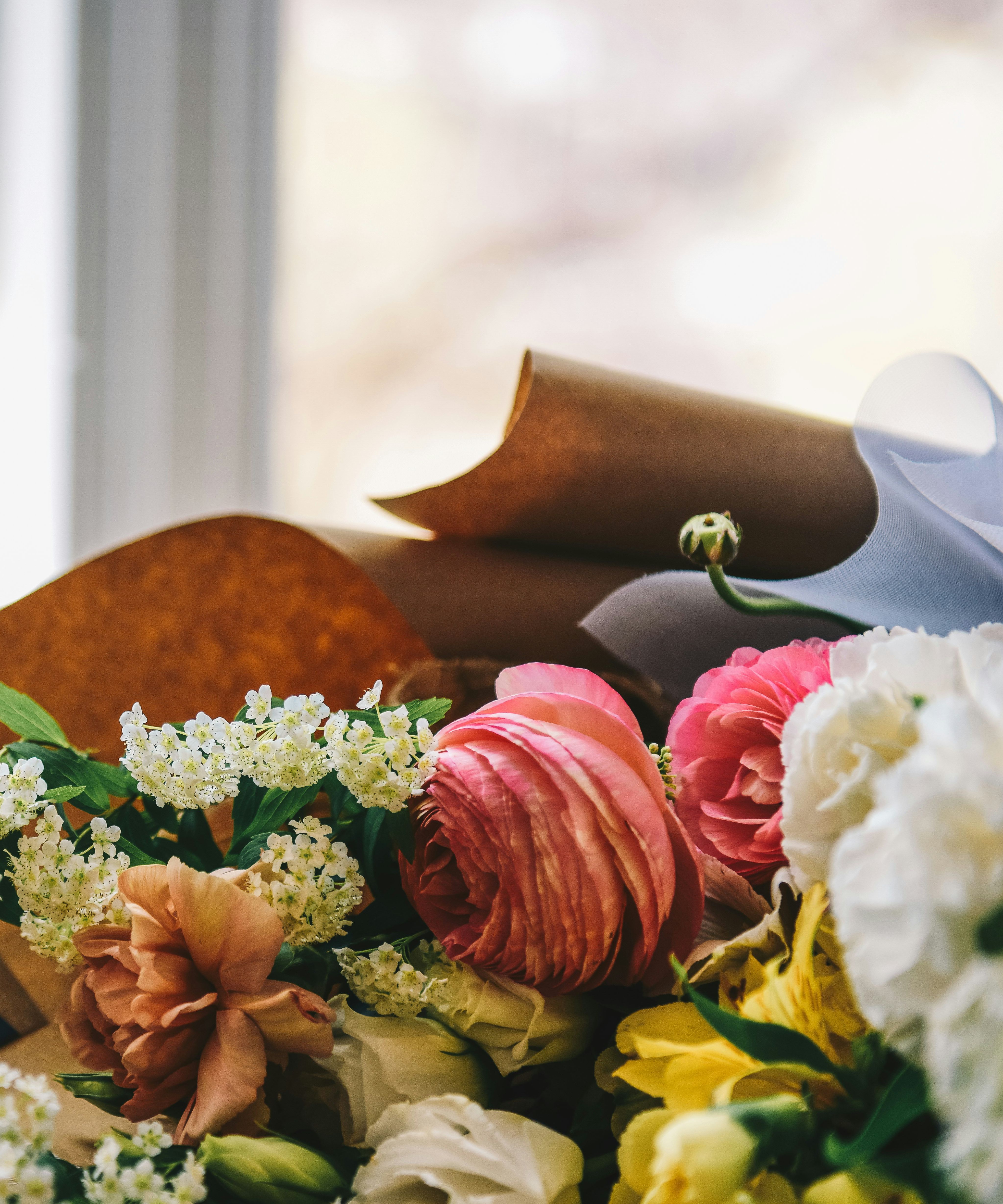 A bouquet of colorful flowers wrapped in brown paper lying on a wooden surface near a window.
