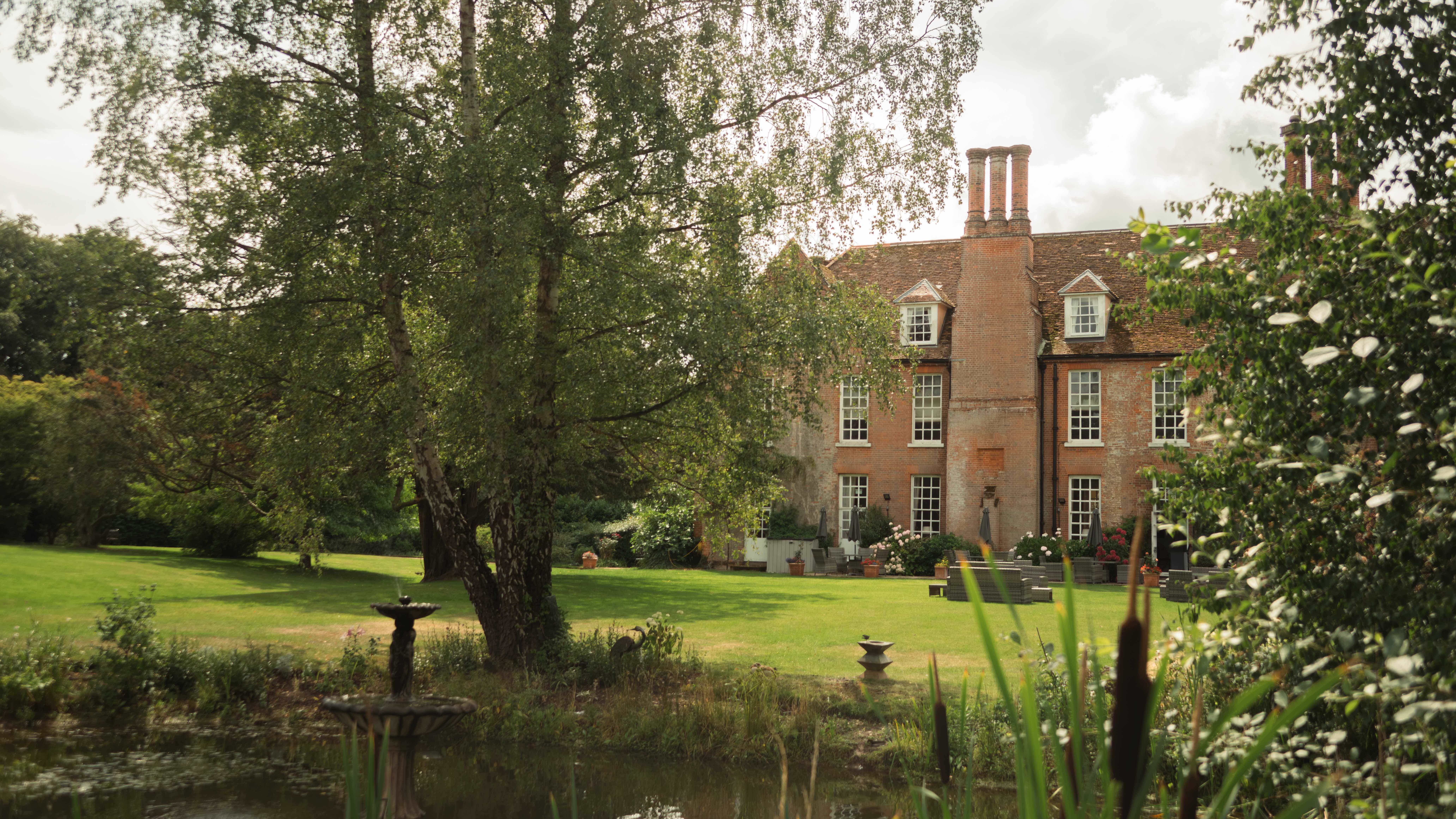 Large brick house seen through trees with a pond and fountain in the foreground