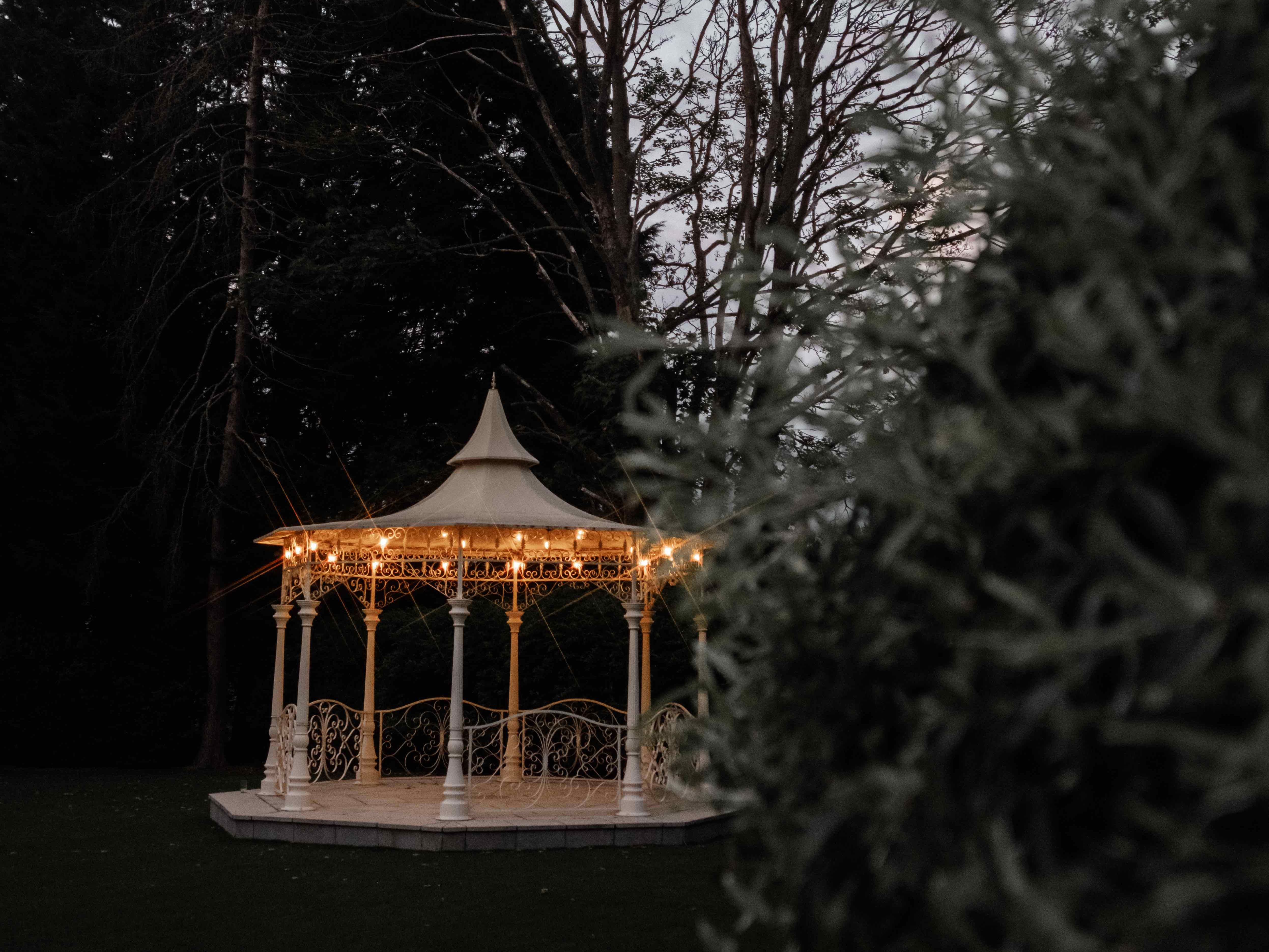 A white gazebo decorated with string lights stands in a garden at dusk, surrounded by trees.