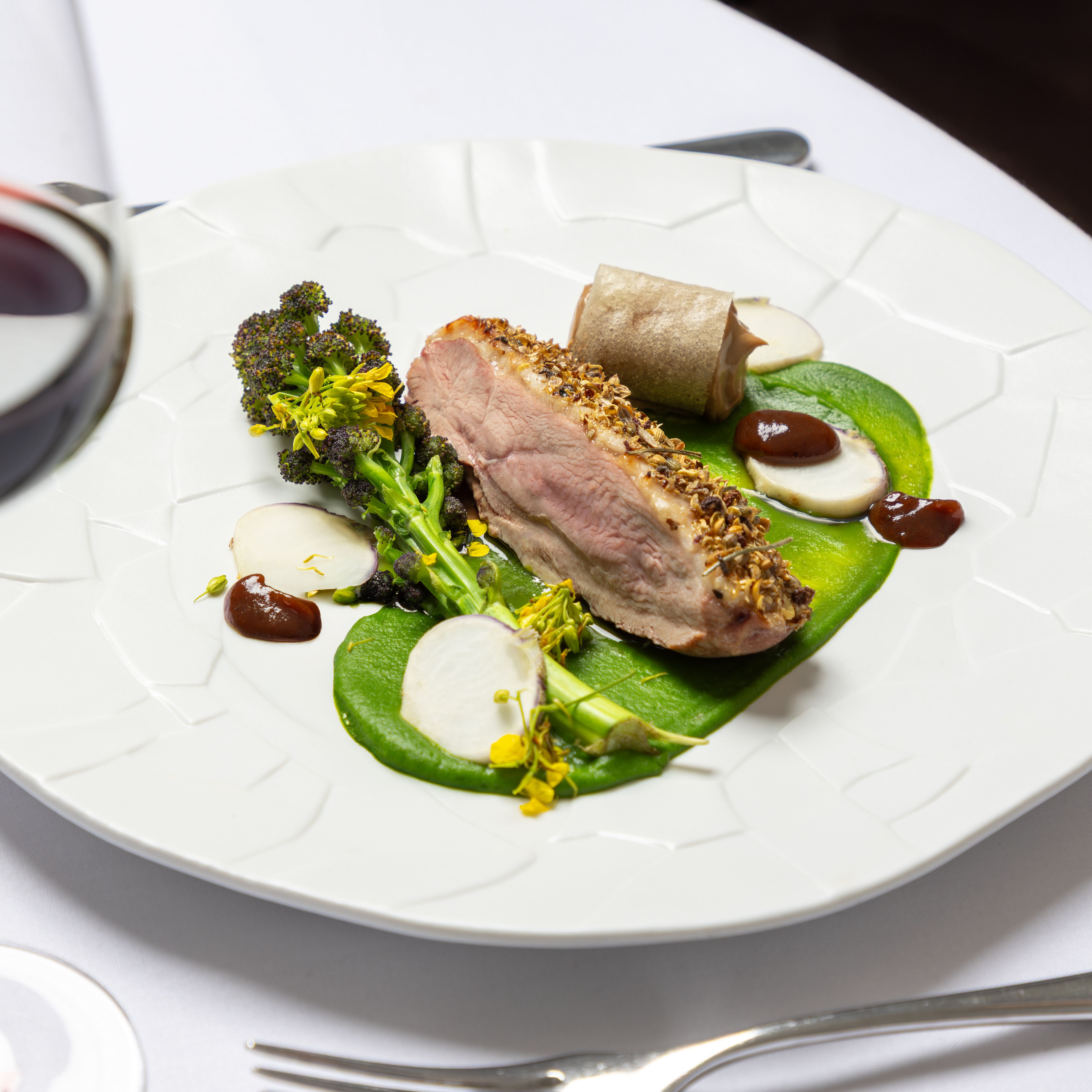 A gourmet dish featuring a sliced piece of seasoned meat, green puree, vegetables, and sauce artfully arranged on a white plate.