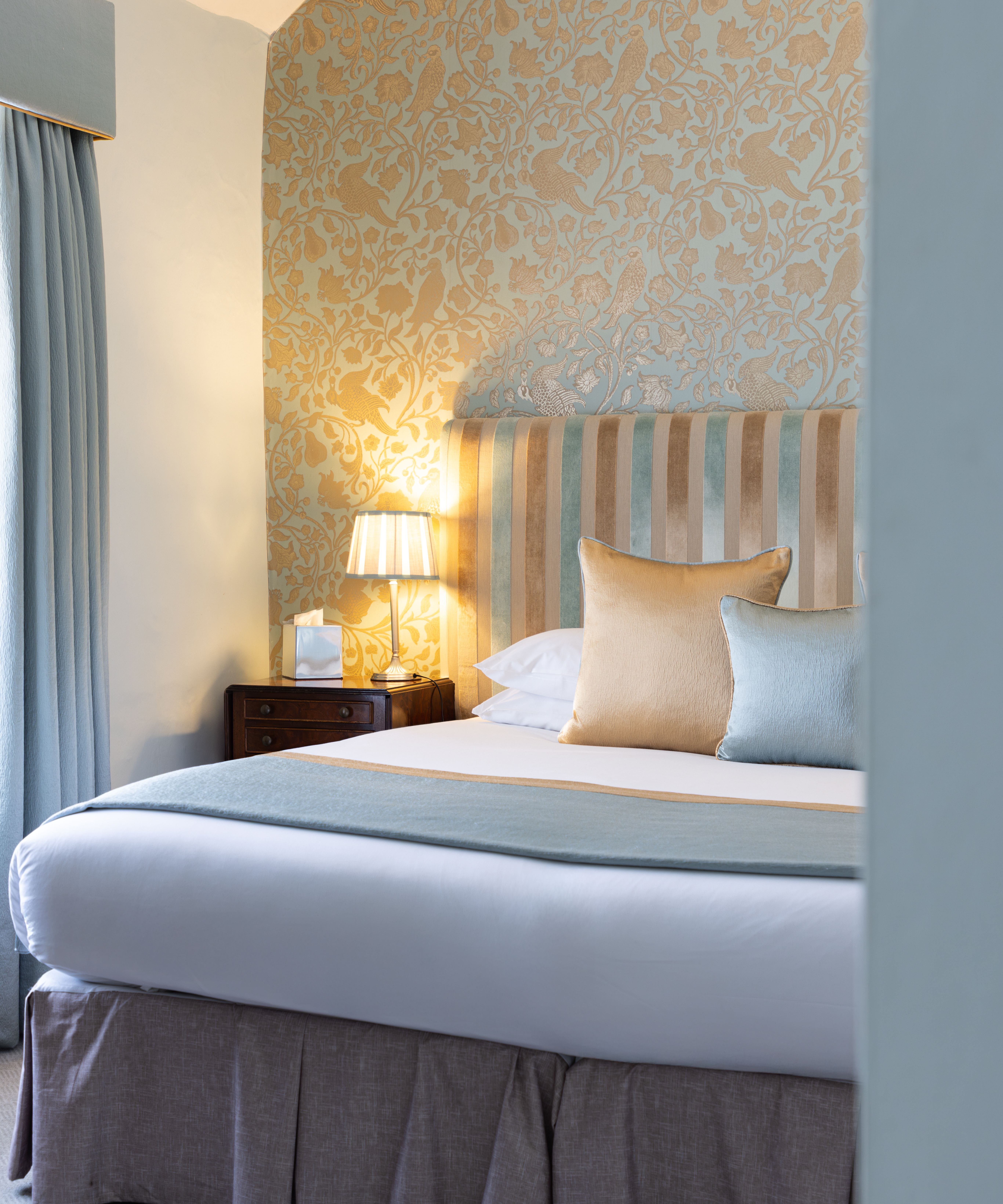 Elegant bedroom with a striped headboard, decorative wallpaper, pillows, bedside table and lamp.
