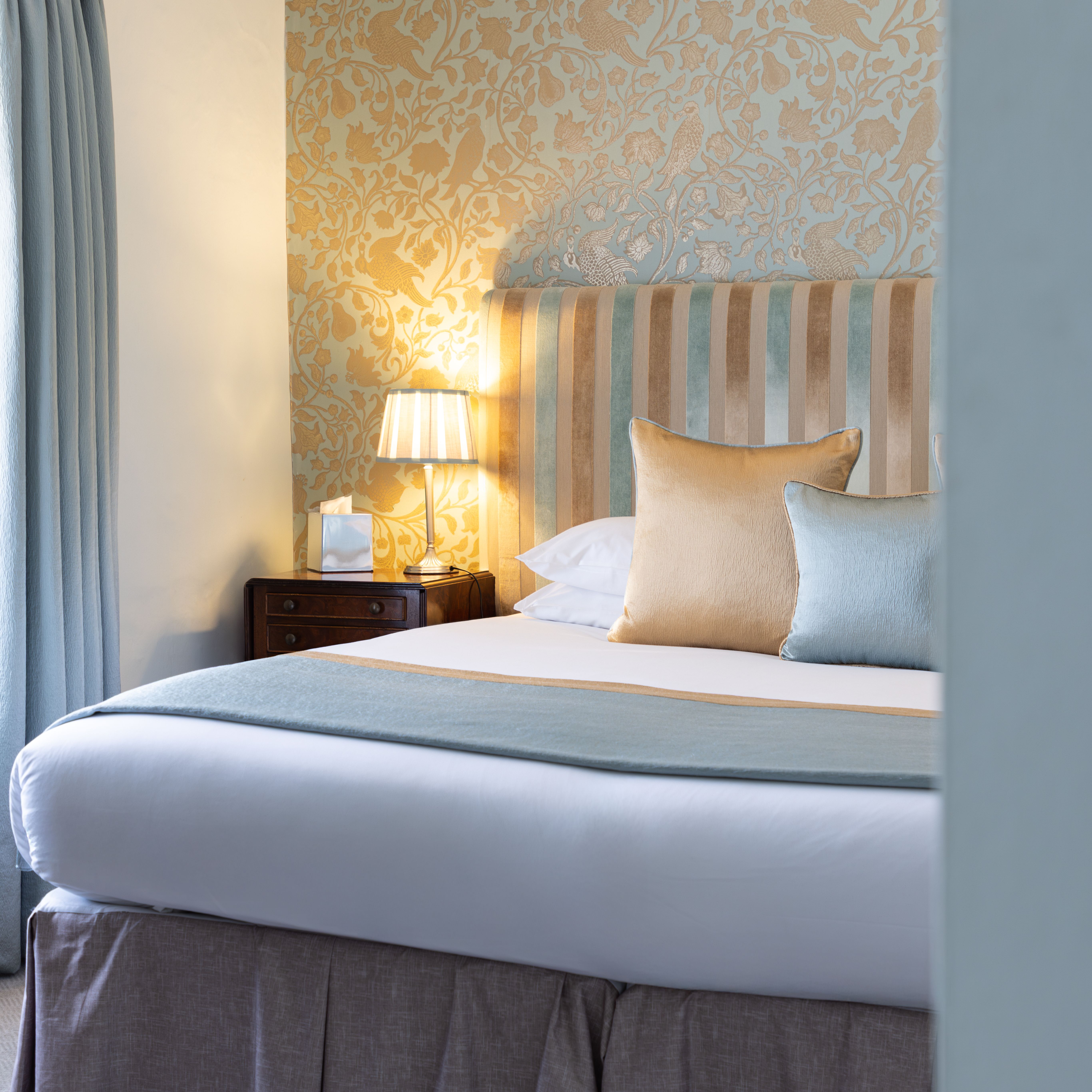 Elegant bedroom with a striped headboard, decorative wallpaper, pillows, bedside table and lamp.