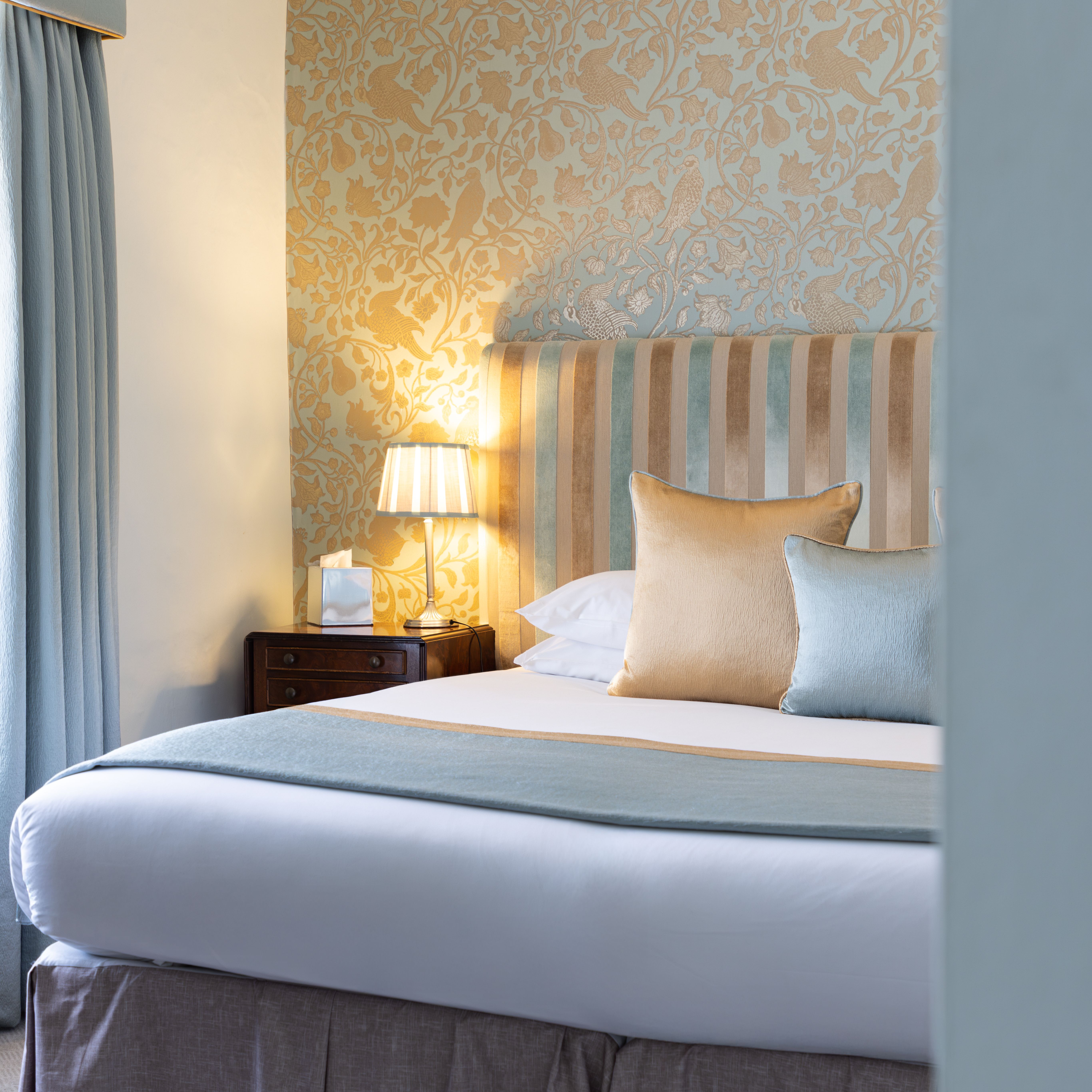 Elegant bedroom with a striped headboard, decorative wallpaper, pillows, bedside table and lamp.