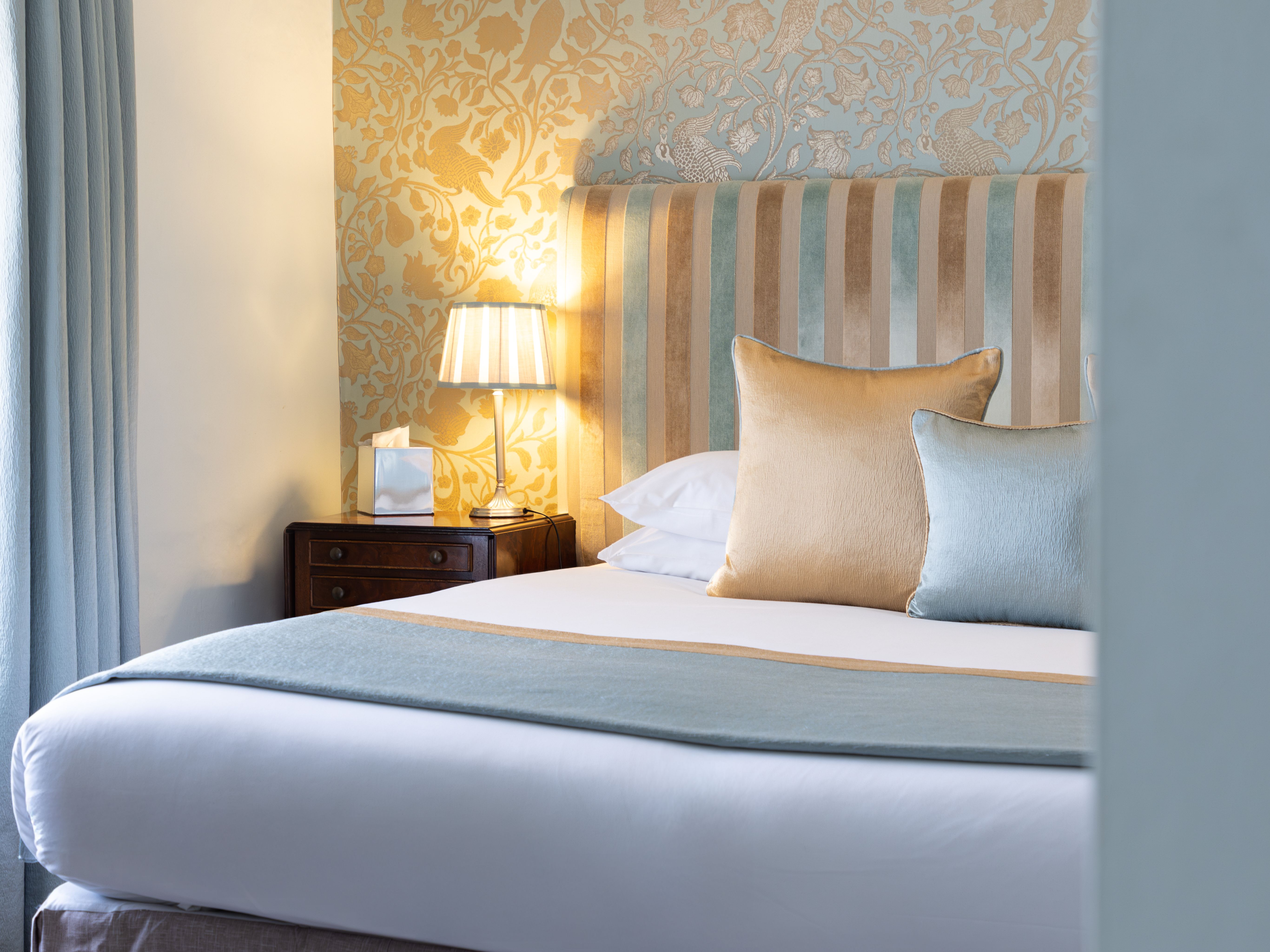 Elegant bedroom with a striped headboard, decorative wallpaper, pillows, bedside table and lamp.