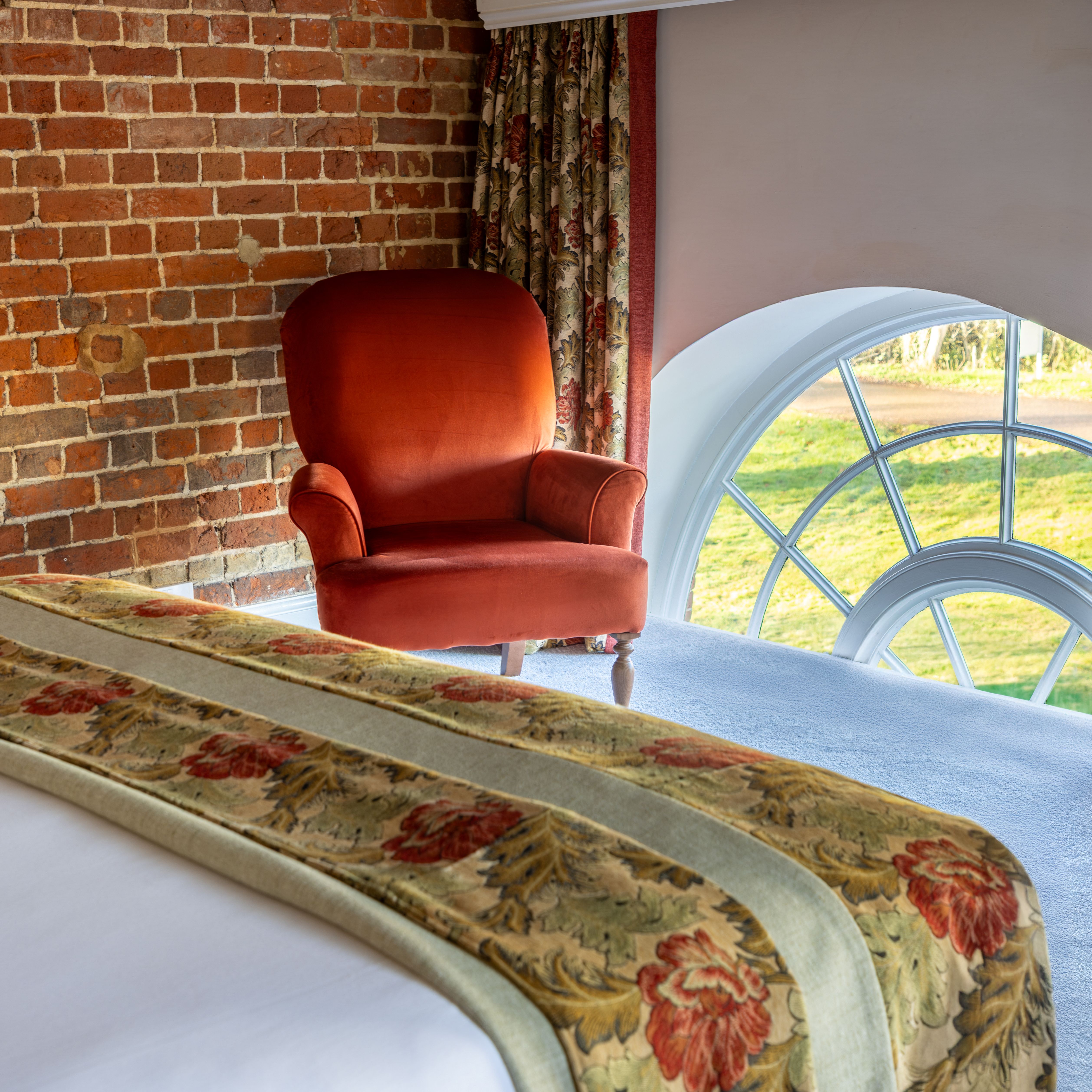 Cozy bedroom with brick wall, red armchair, floral curtains, and arched window.