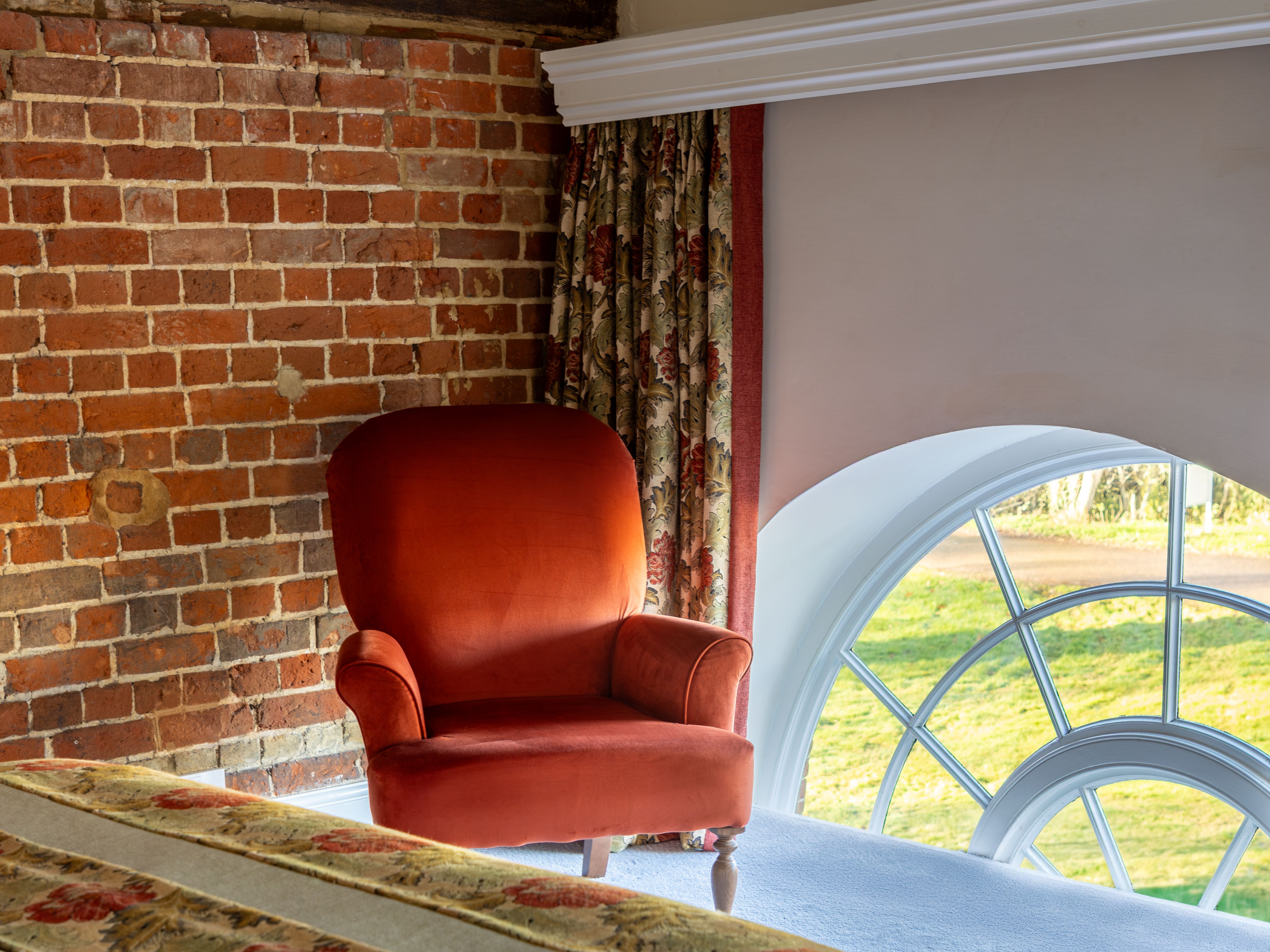 Cozy bedroom with brick wall, red armchair, floral curtains, and arched window.