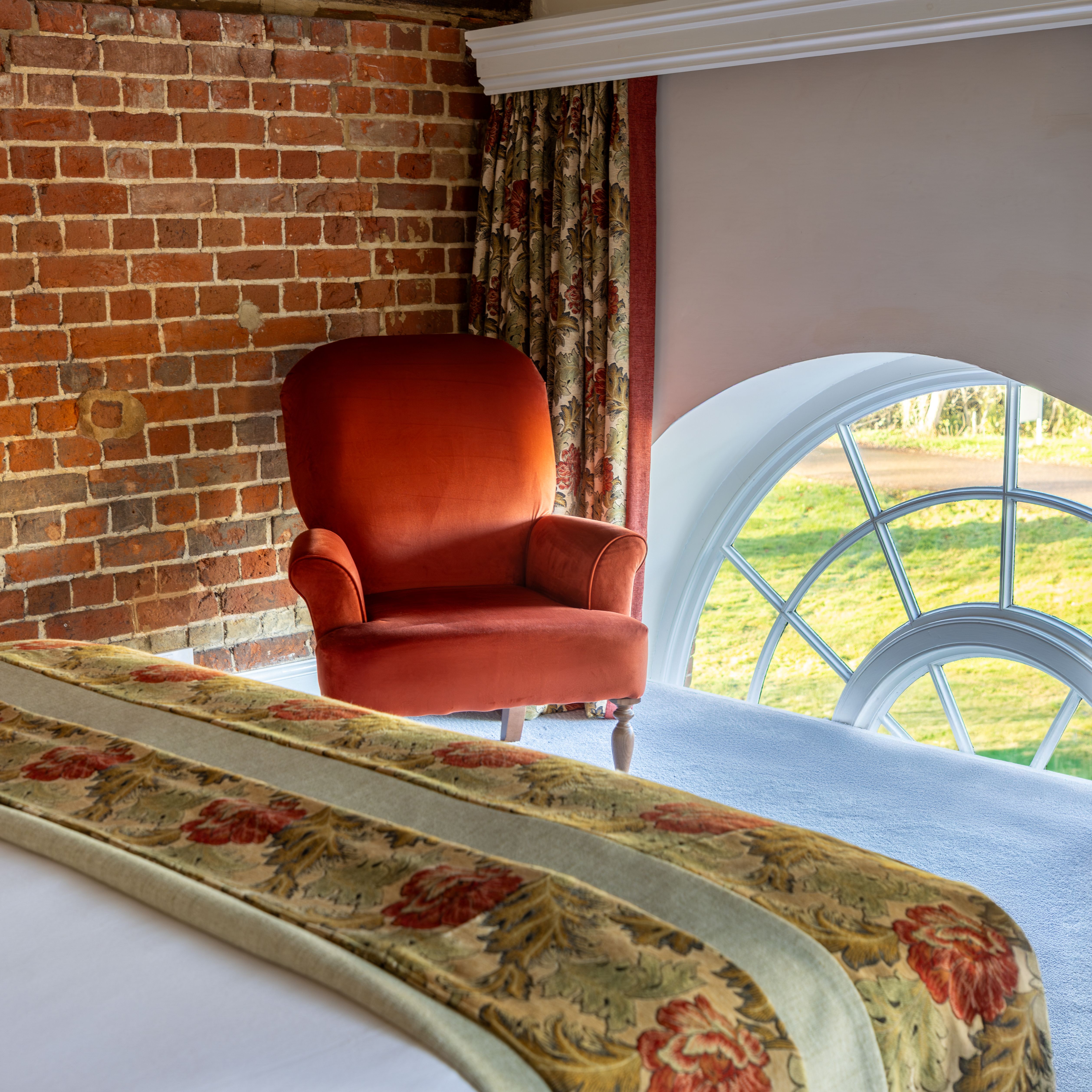 Cozy bedroom with brick wall, red armchair, floral curtains, and arched window.