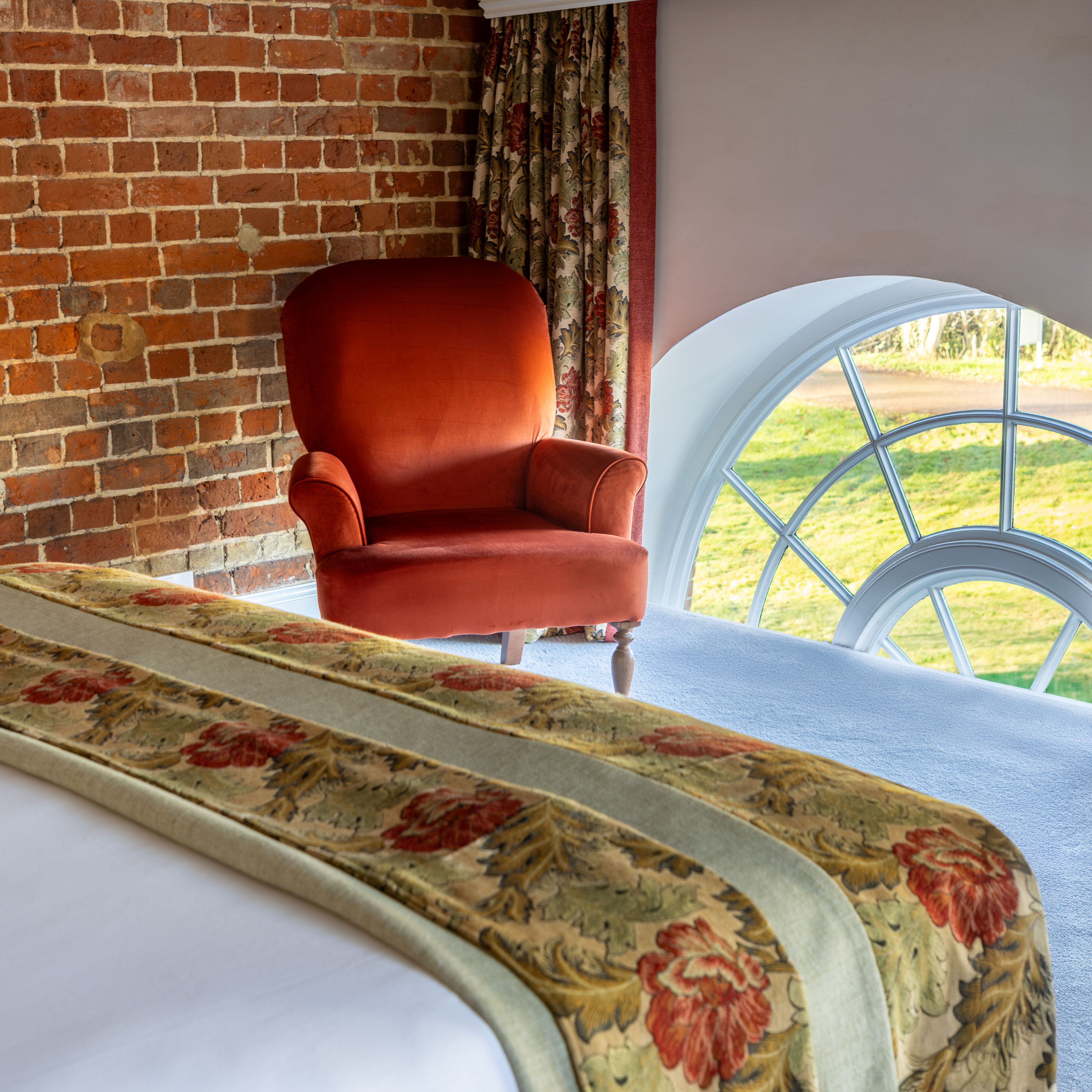 Cozy bedroom with brick wall, red armchair, floral curtains, and arched window.