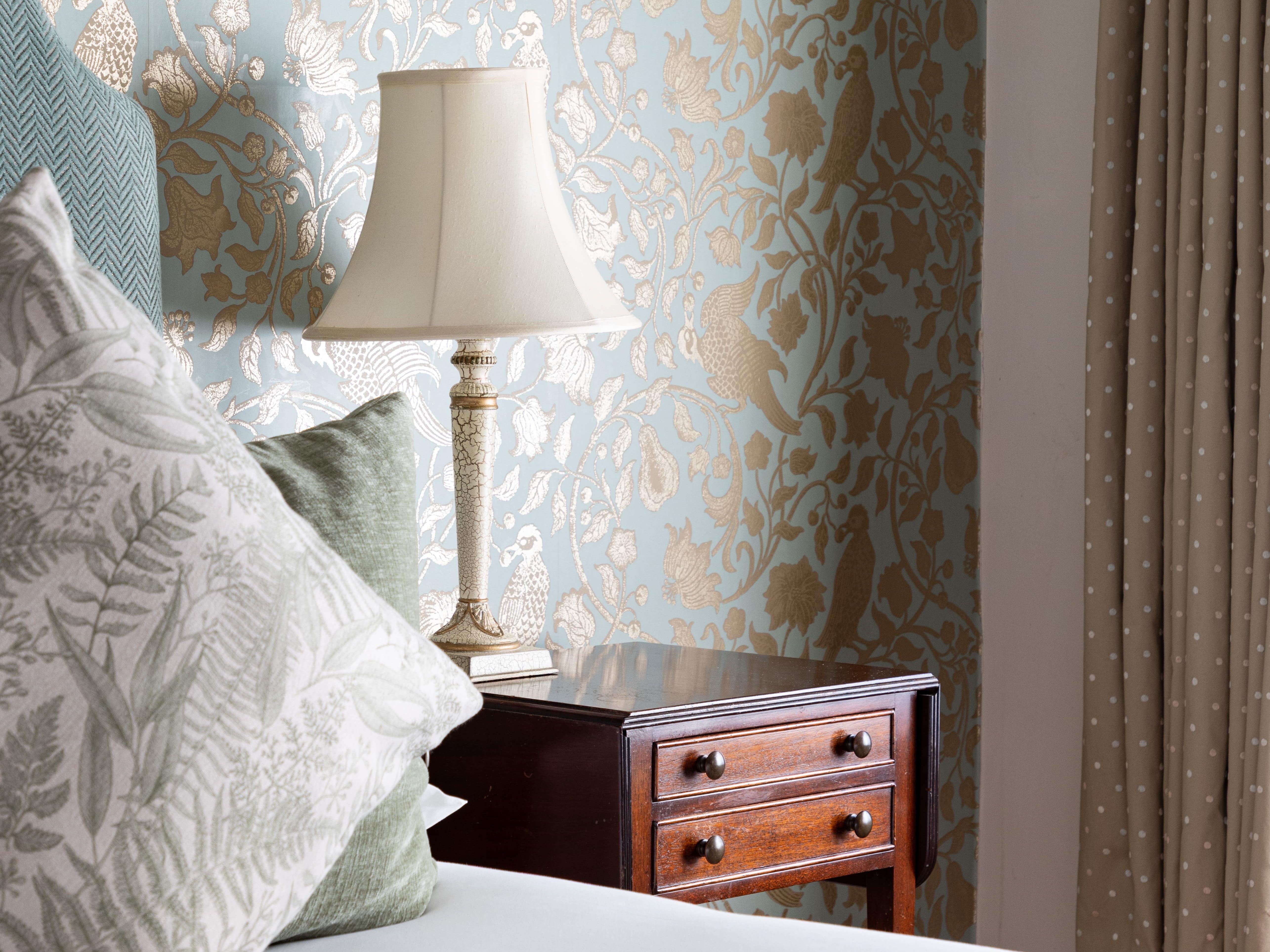 Bedroom corner with patterned wallpaper, bedside table, lamp, and pillows