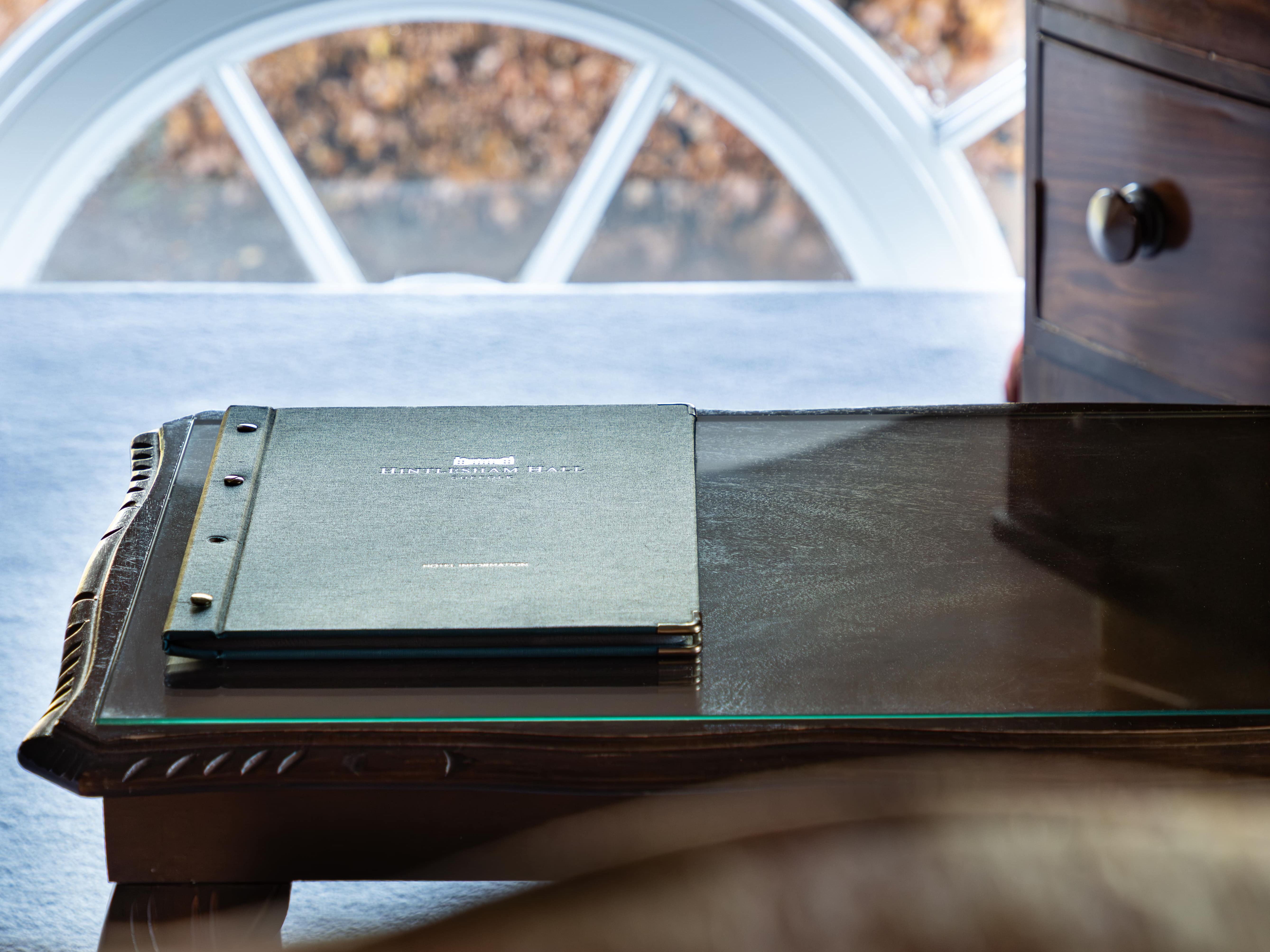 A close-up of a dark wooden desk with a green notebook on it, next to a window with an arched frame.
