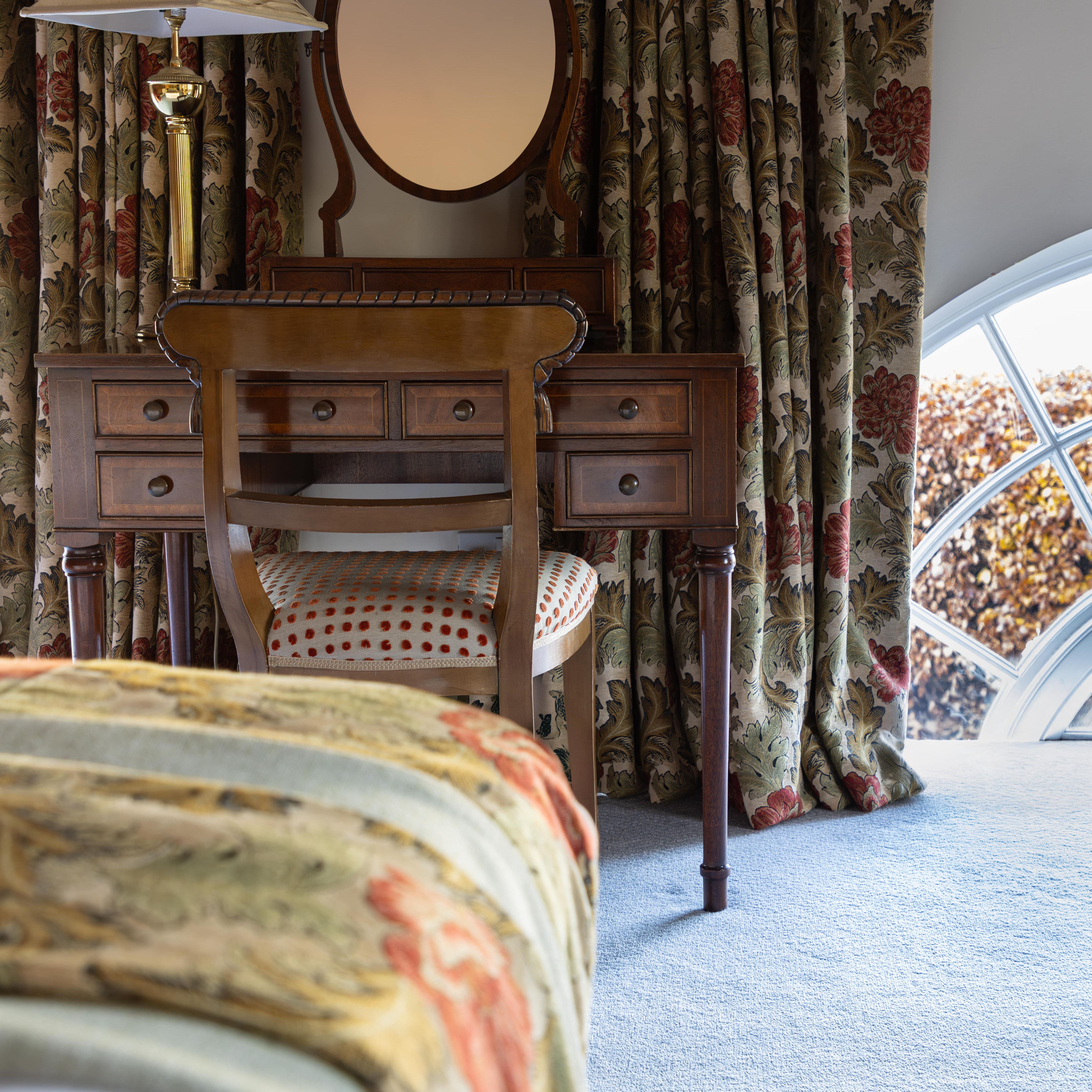 Traditional bedroom with floral curtains, wooden vanity, chair, and lamp