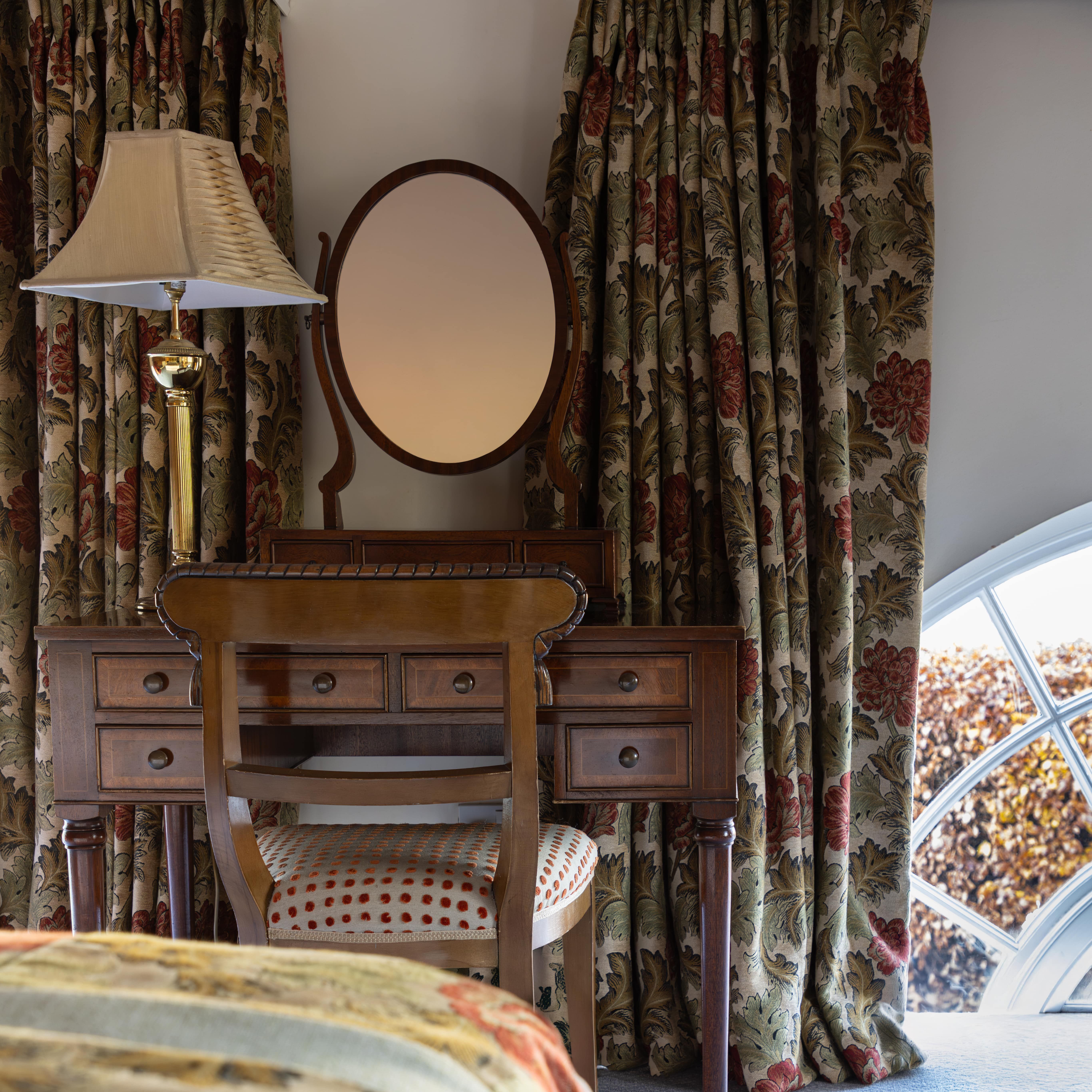 Traditional bedroom with floral curtains, wooden vanity, chair, and lamp