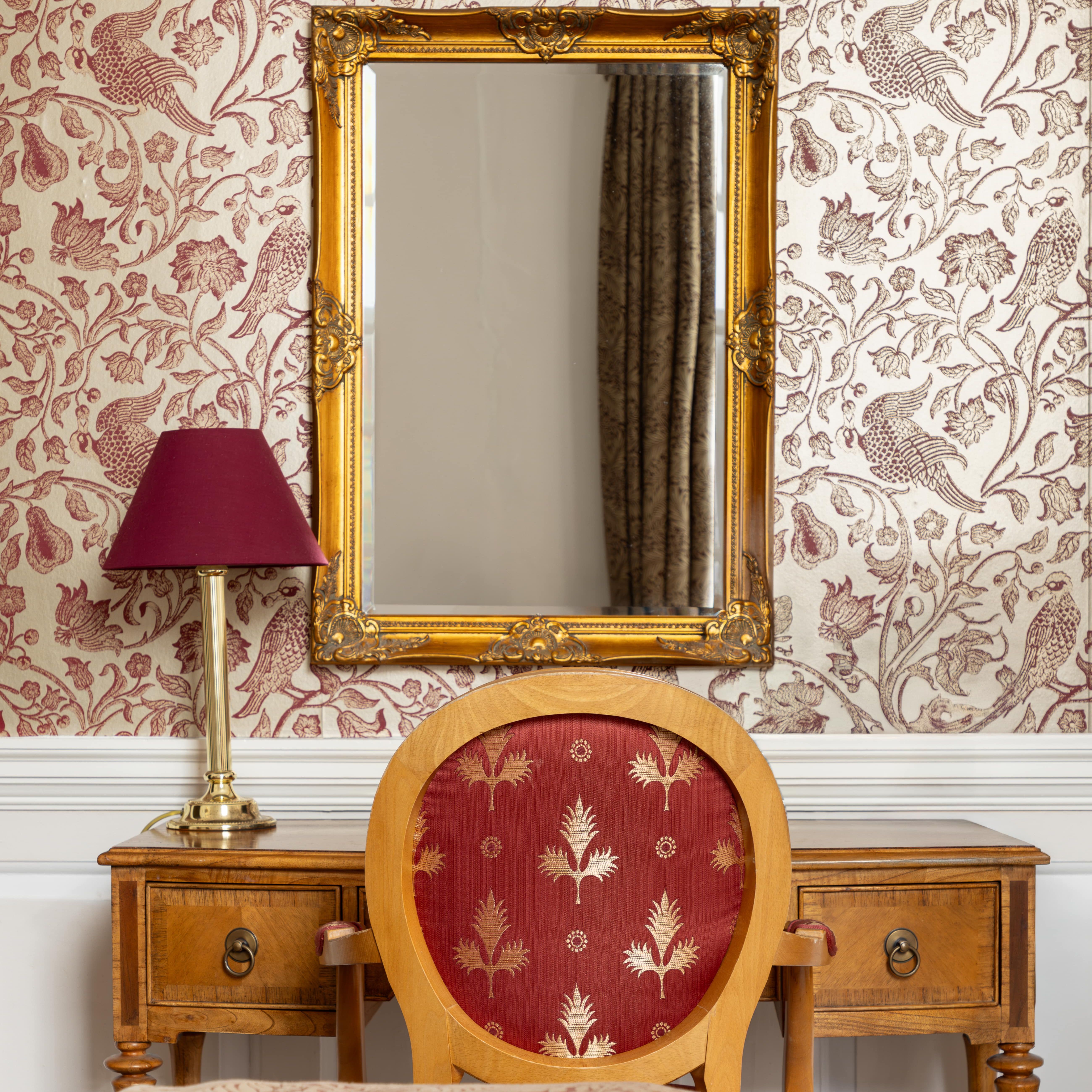 Elegant bedroom with a vintage wooden desk, ornate gold-framed mirror, and red-patterned chair