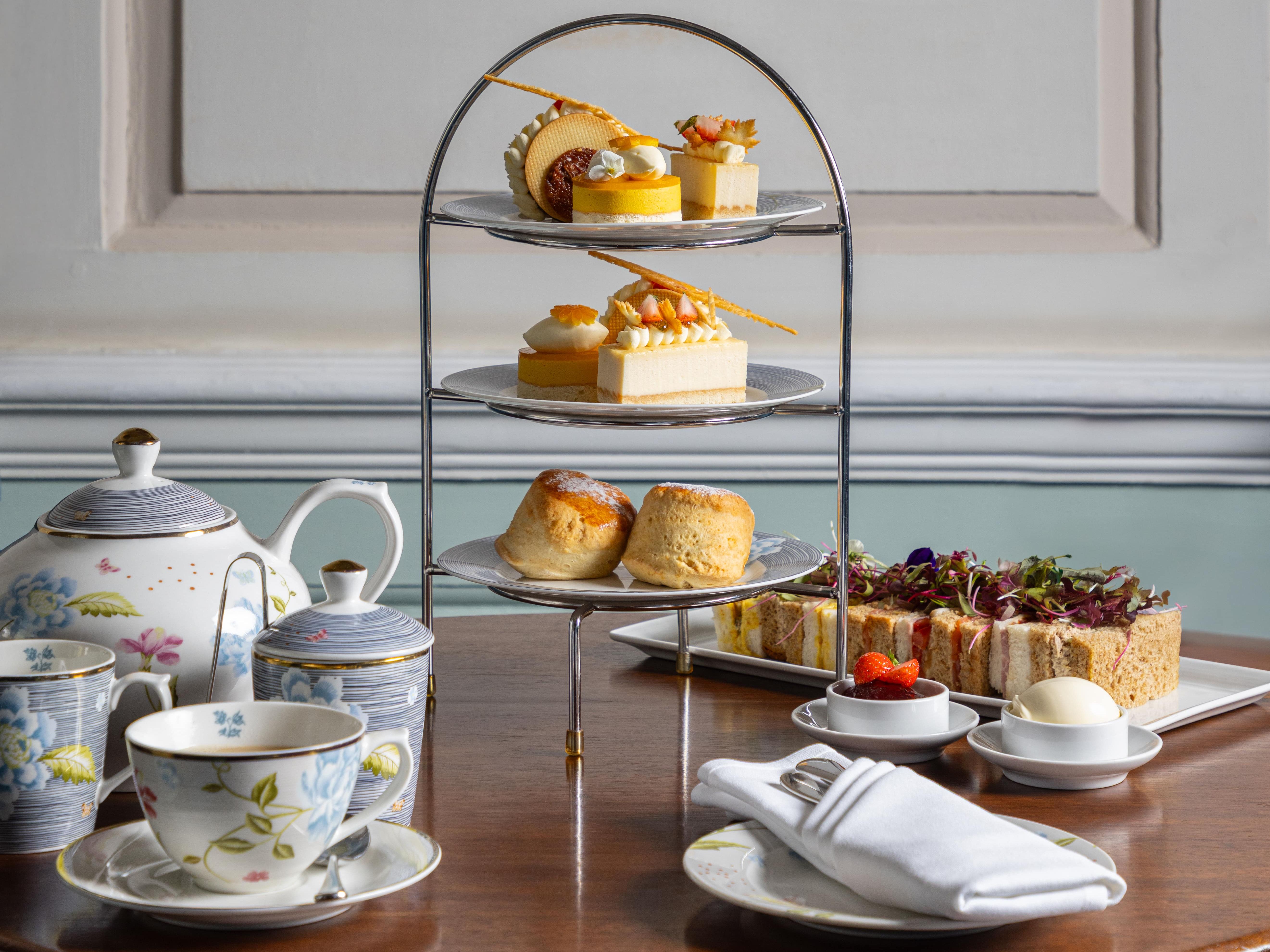 Elegant afternoon tea setup with a tiered tray of pastries, scones, and desserts, accompanied by a teapot, cups, and plates of finger sandwiches.