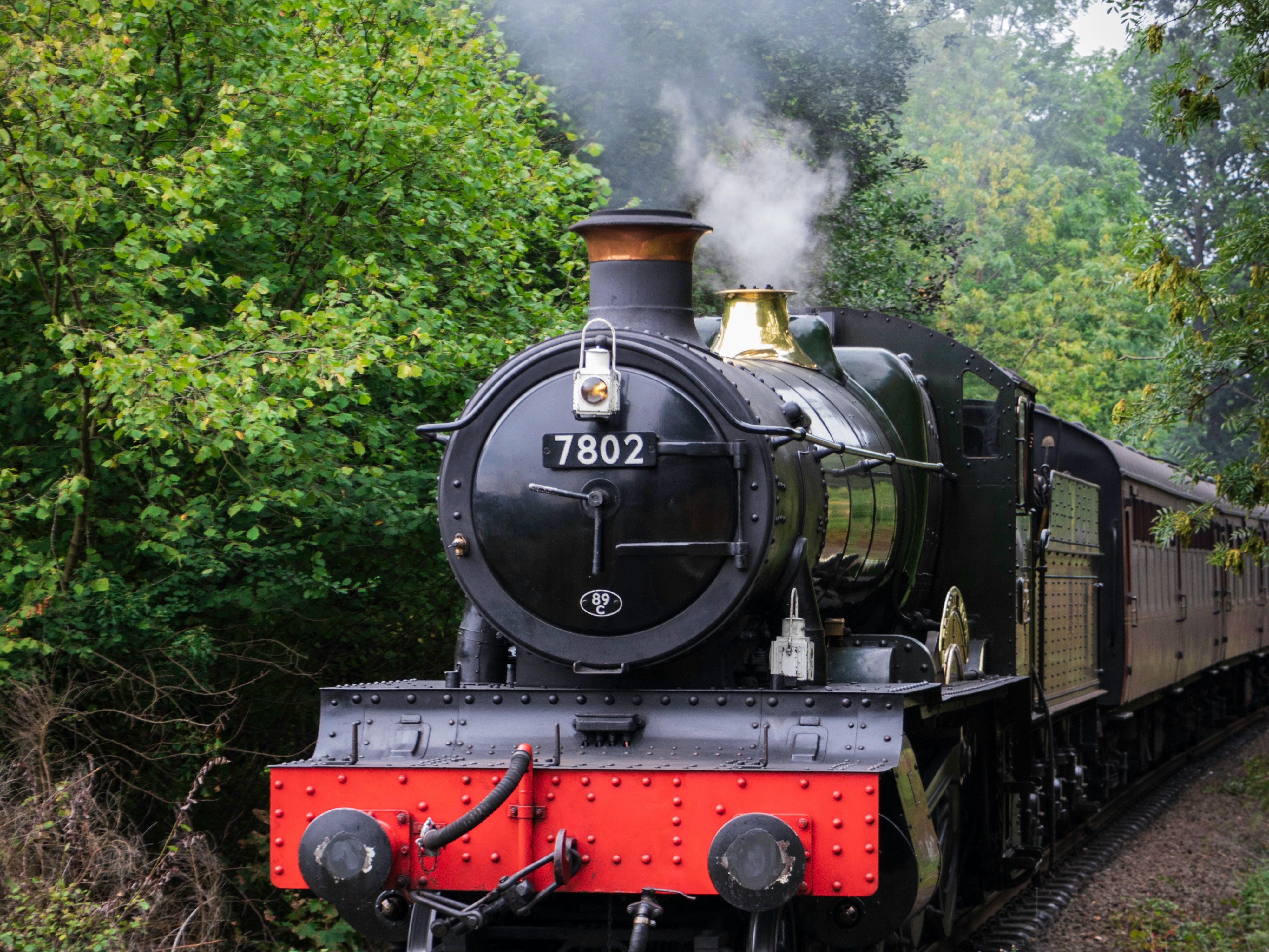 Vintage steam locomotive train traveling on railway tracks through a green wooded area.