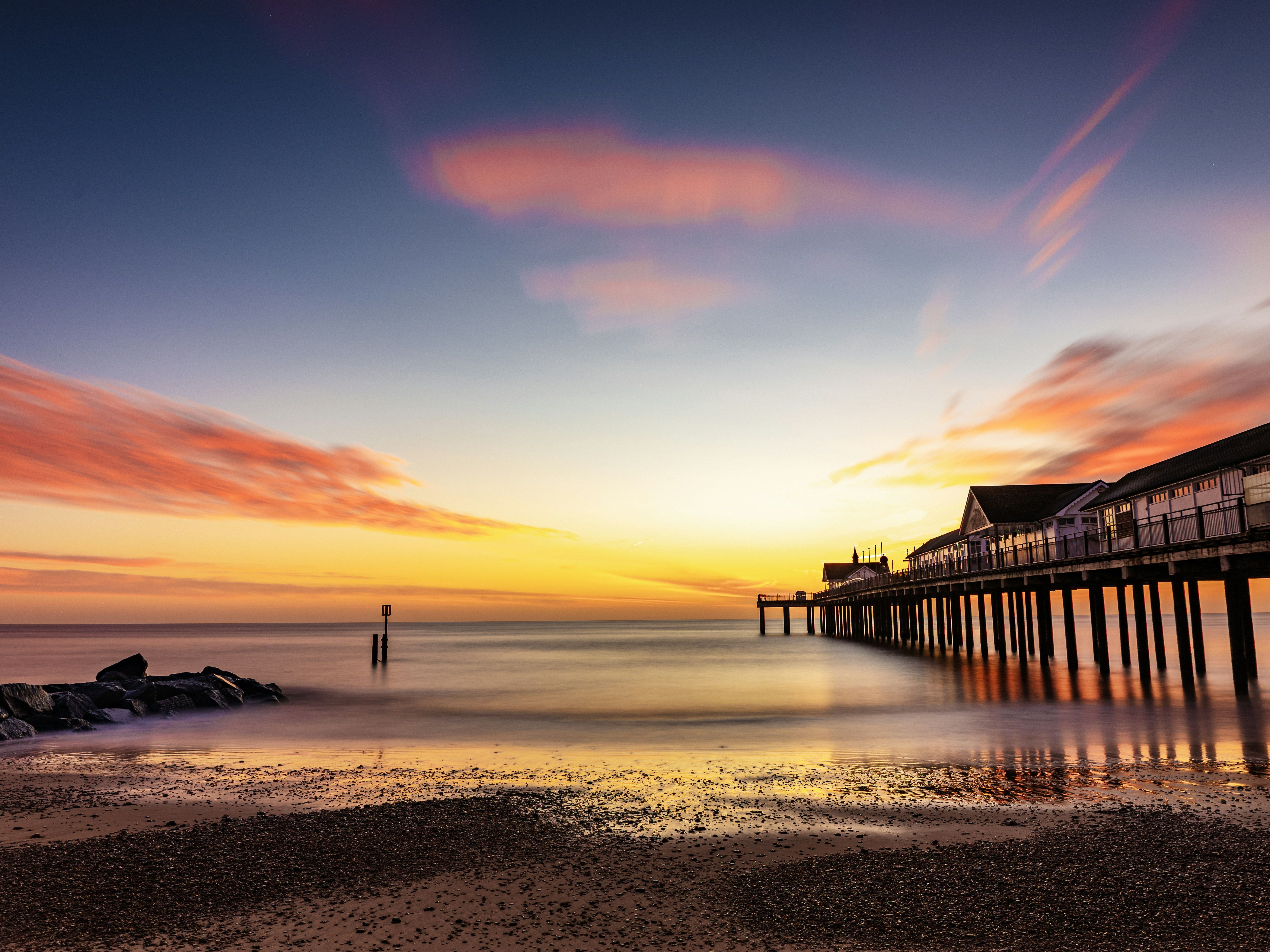 Pier stretching over calm water at sunset with dramatic clouds and reflections.