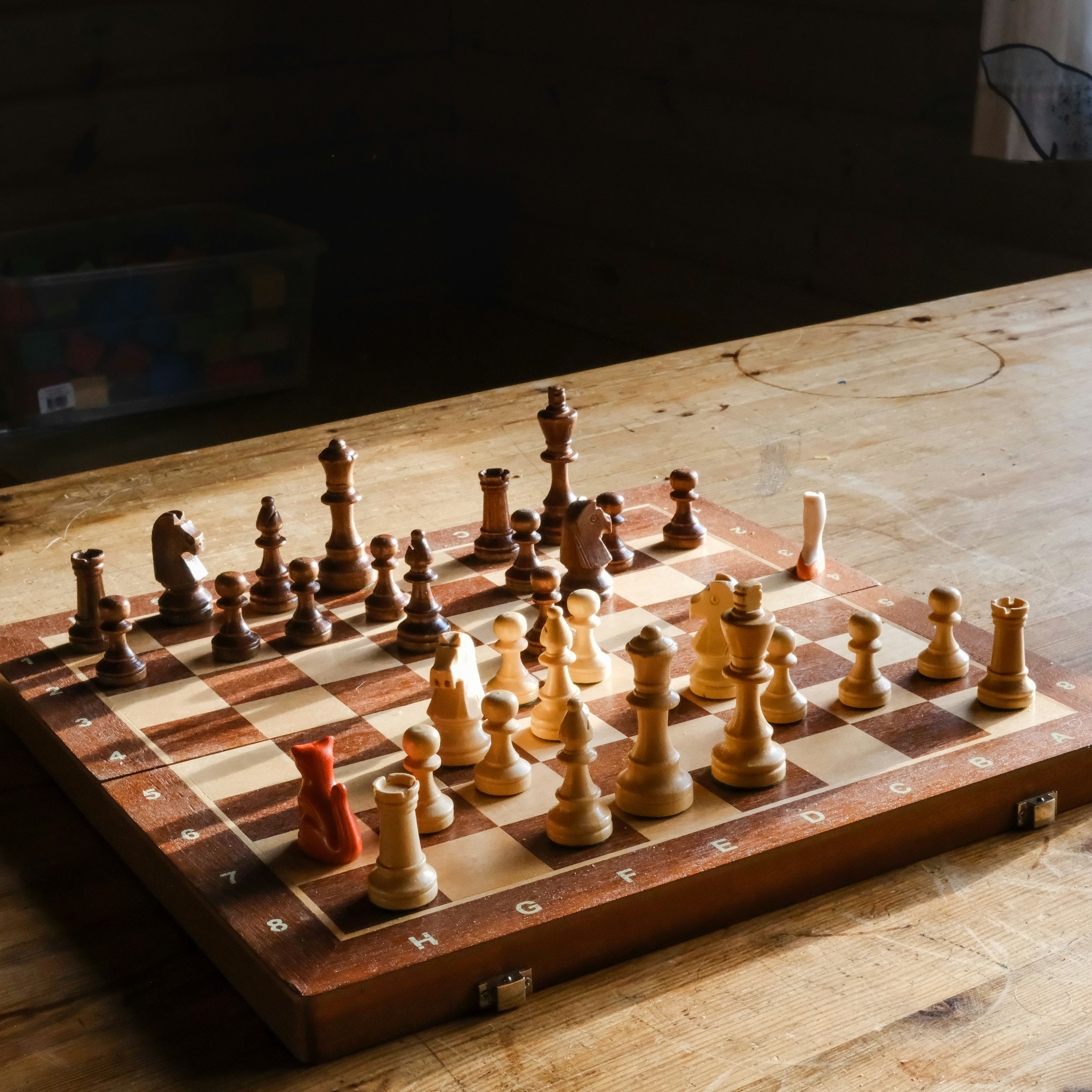 A wooden chessboard with chess pieces set up on a wooden table in a sunlit room.