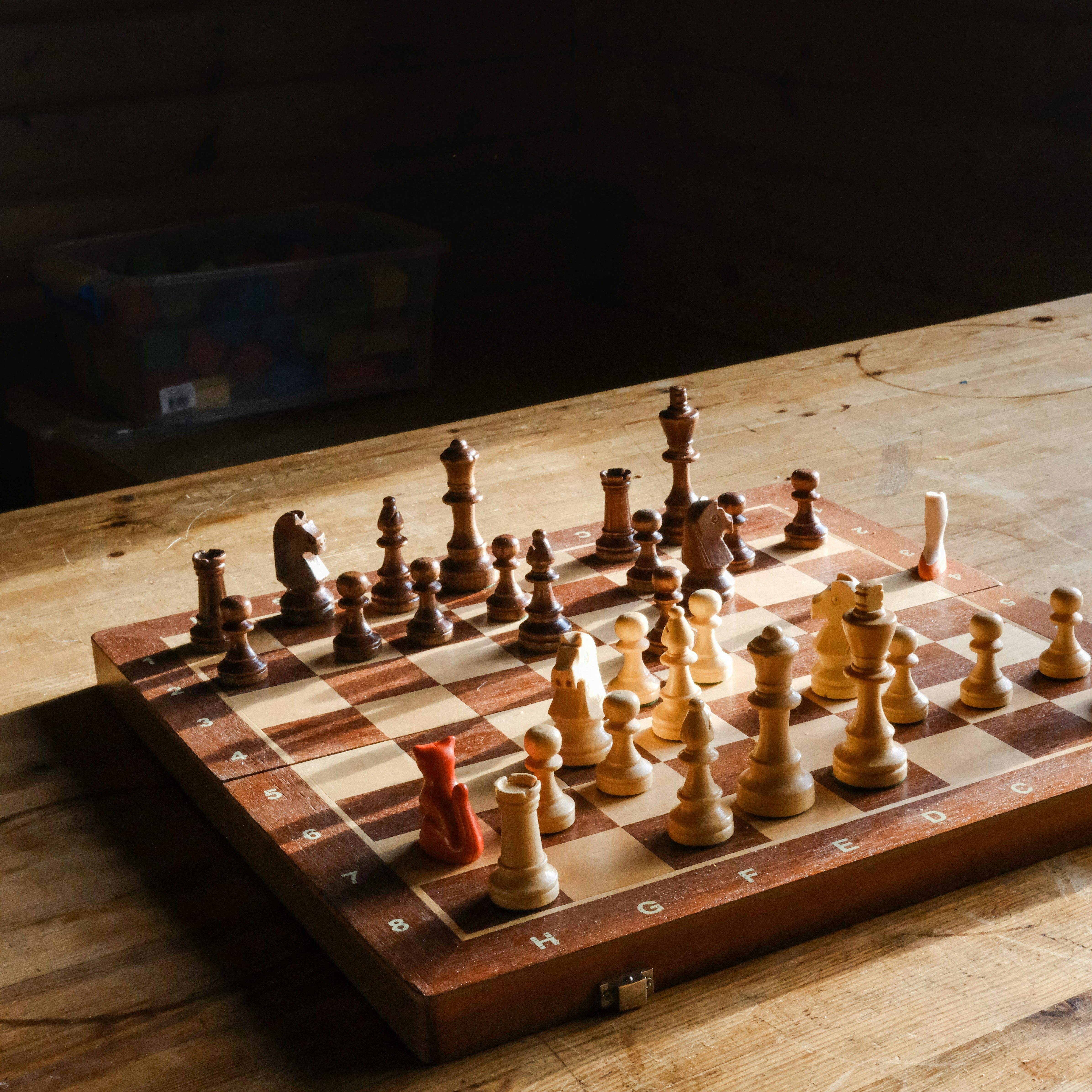 A wooden chessboard with chess pieces set up on a wooden table in a sunlit room.