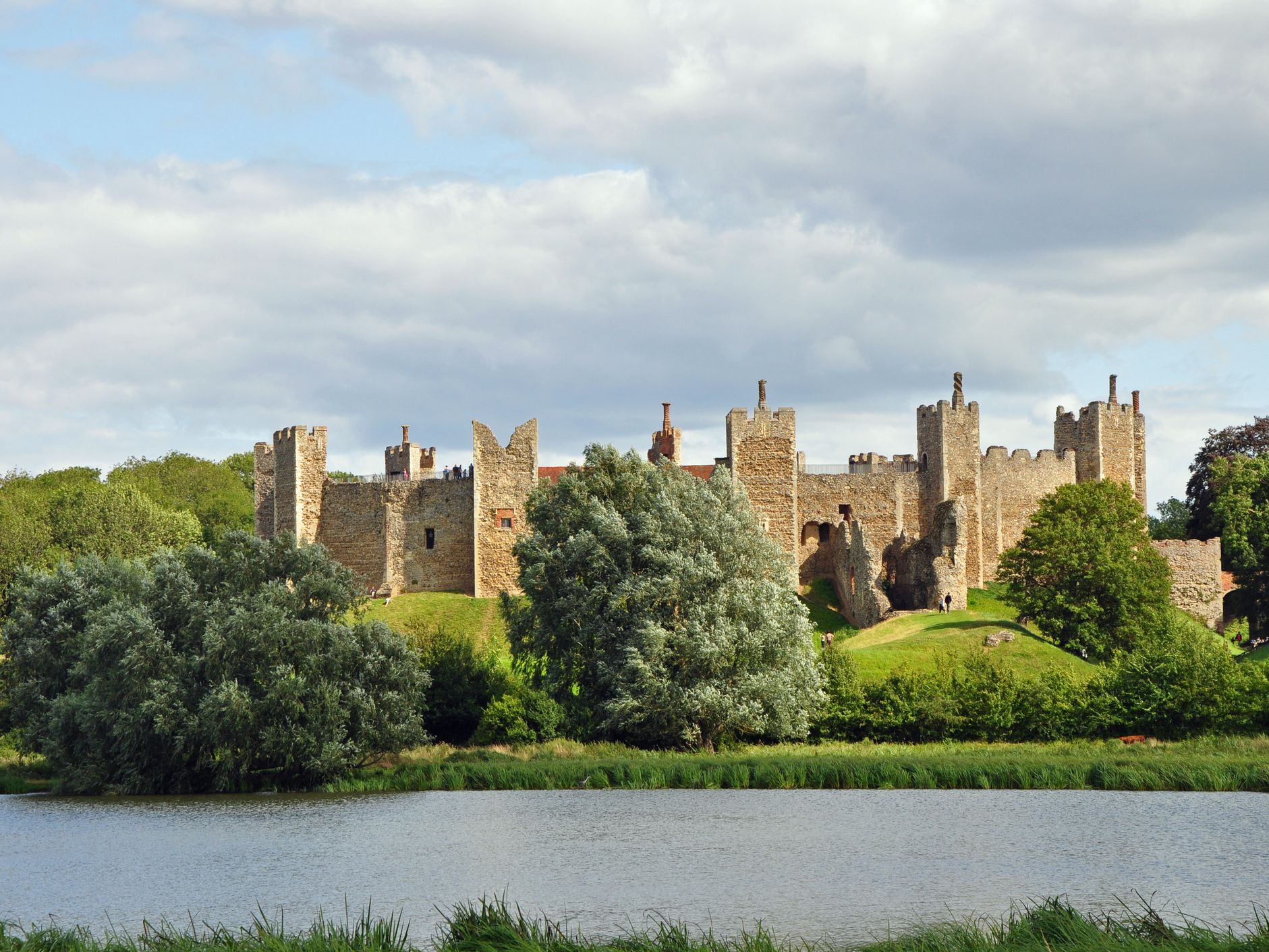 Medieval stone castle with towers and battlements surrounded by trees and a river in the foreground