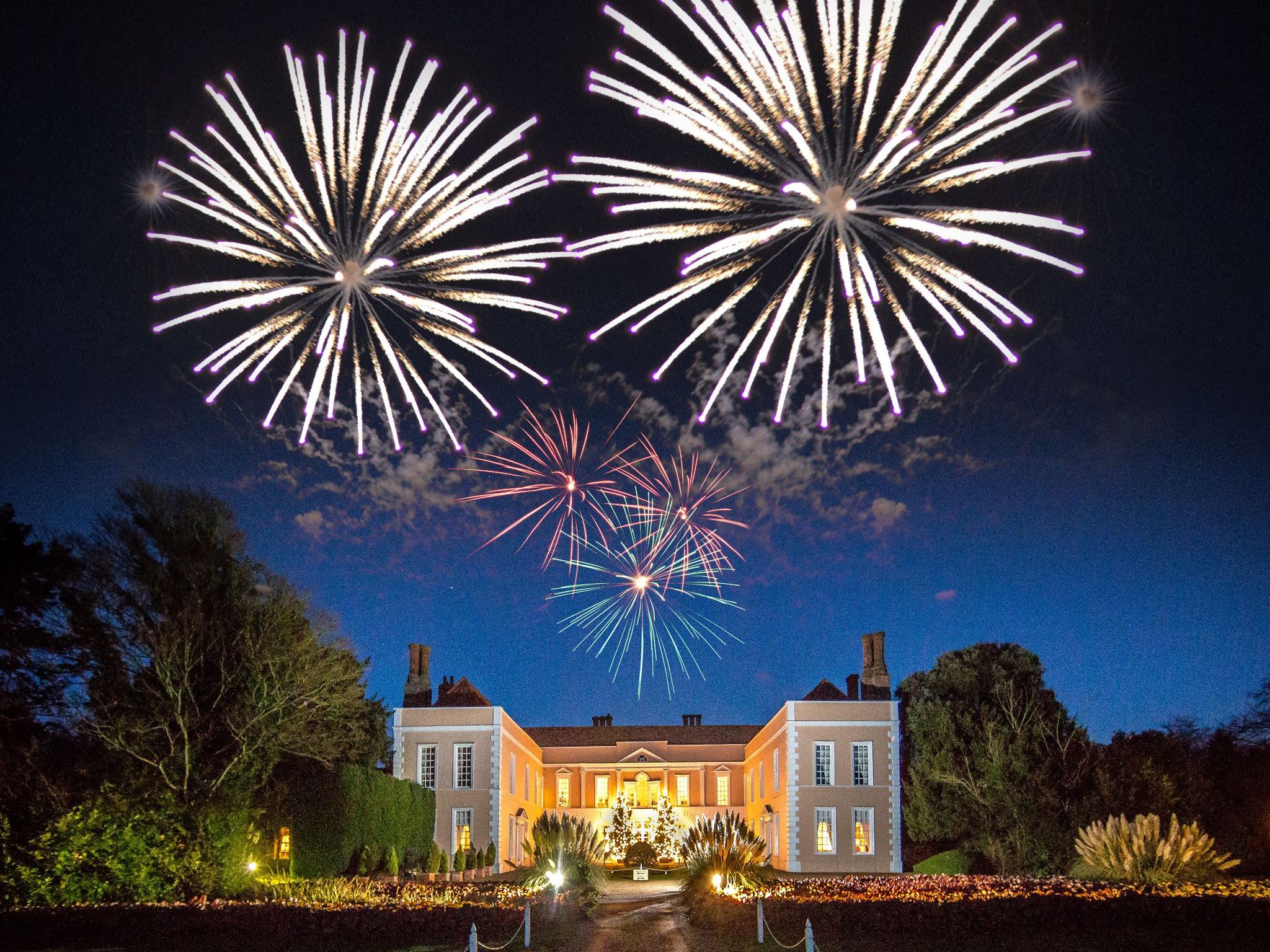 Large manor house illuminated at night with colorful fireworks lighting up the sky above.