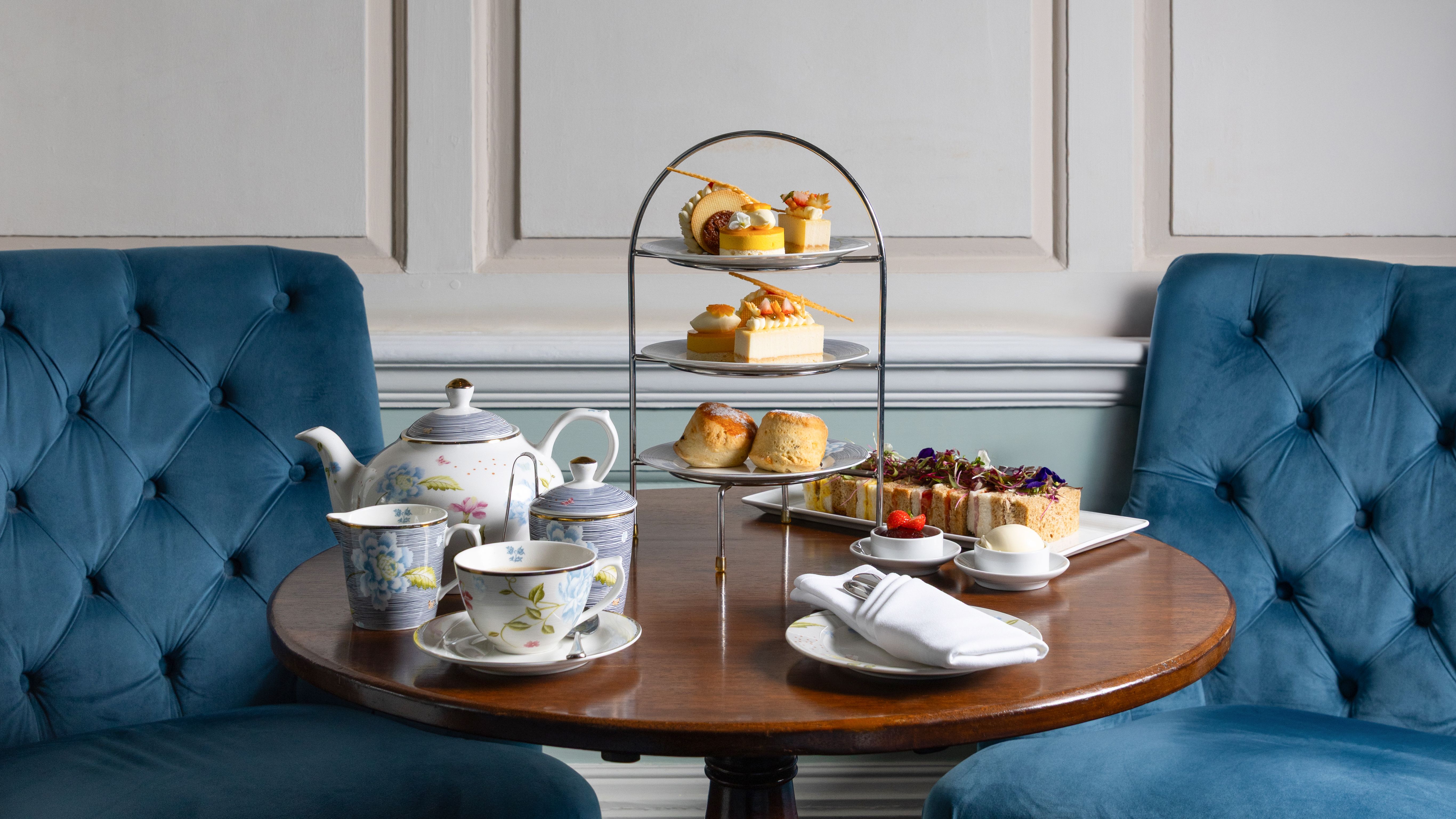 Elegant afternoon tea setup with pastries, scones, tea set, and sandwiches on a round wooden table between two blue velvet chairs.