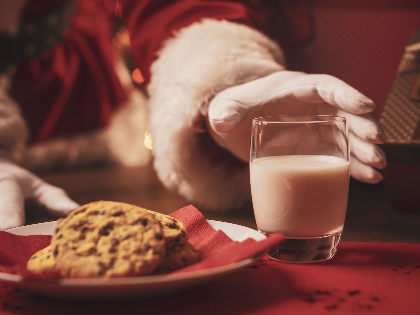 Santa Claus reaching for a glass of milk next to a plate of chocolate chip cookies with a Christmas tree in the background.