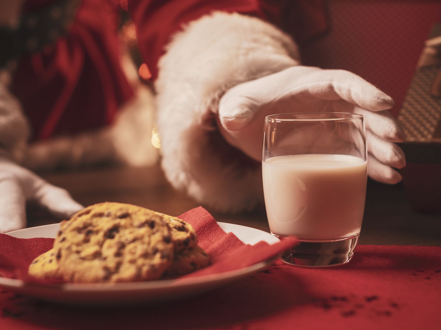 Santa Claus reaching for a glass of milk next to a plate of chocolate chip cookies with a Christmas tree in the background.