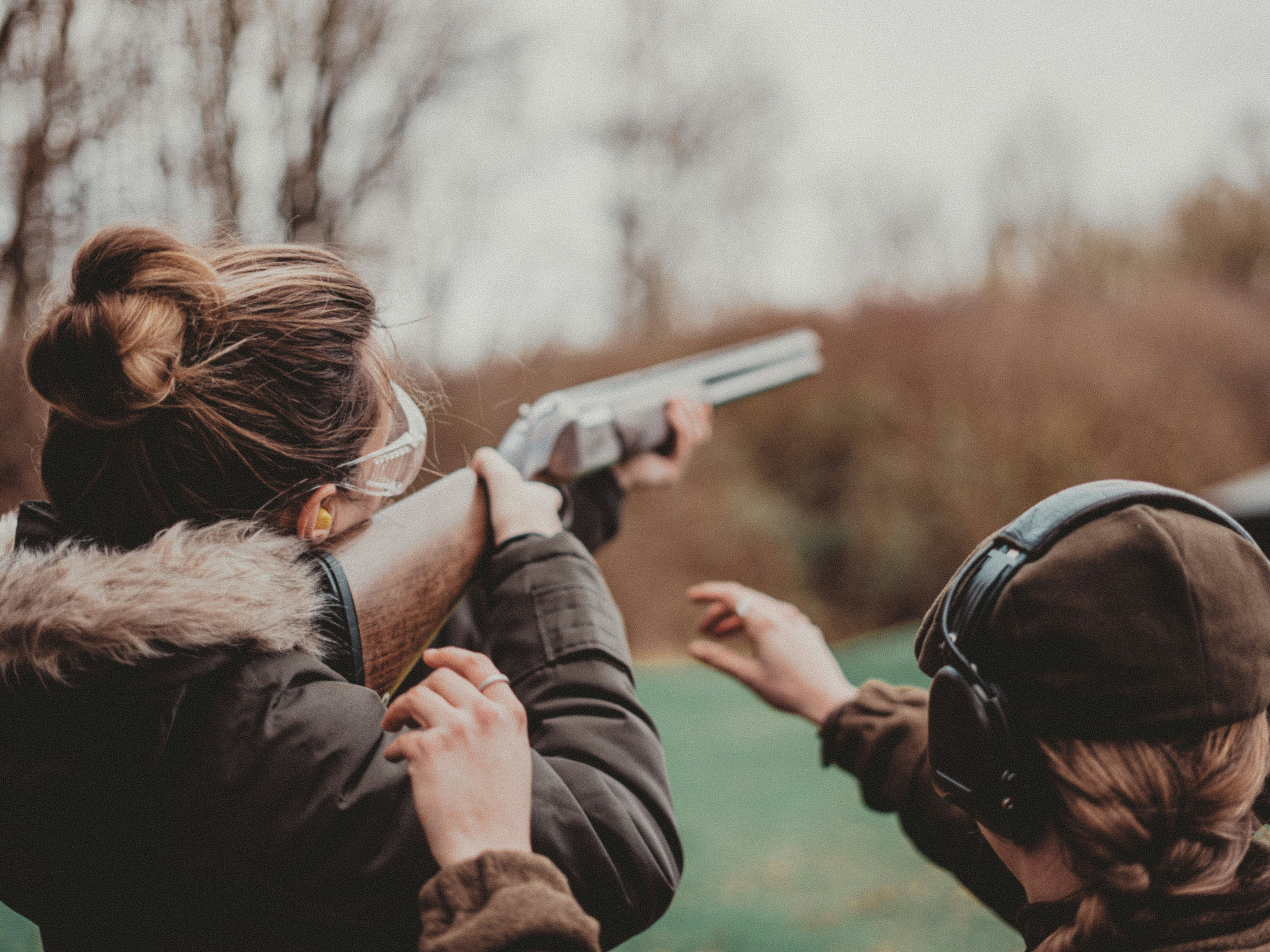 Two people outdoors, one aiming a shotgun and the other providing guidance, both wearing protective gear