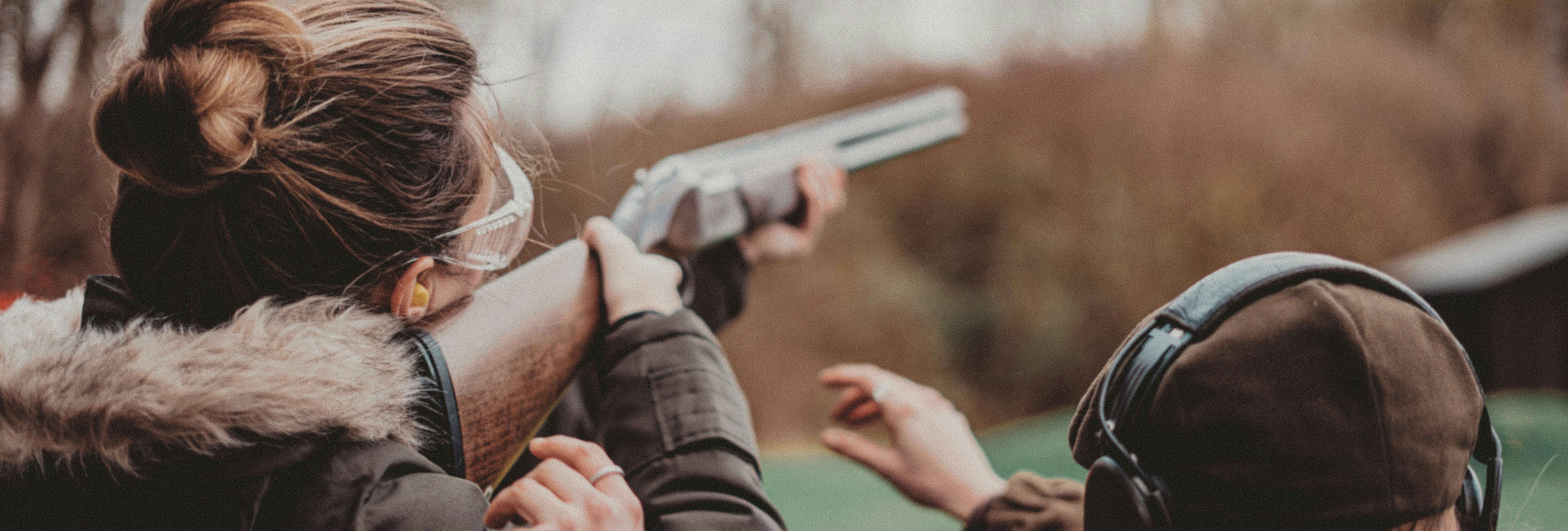 Two people outdoors, one aiming a shotgun and the other providing guidance, both wearing protective gear