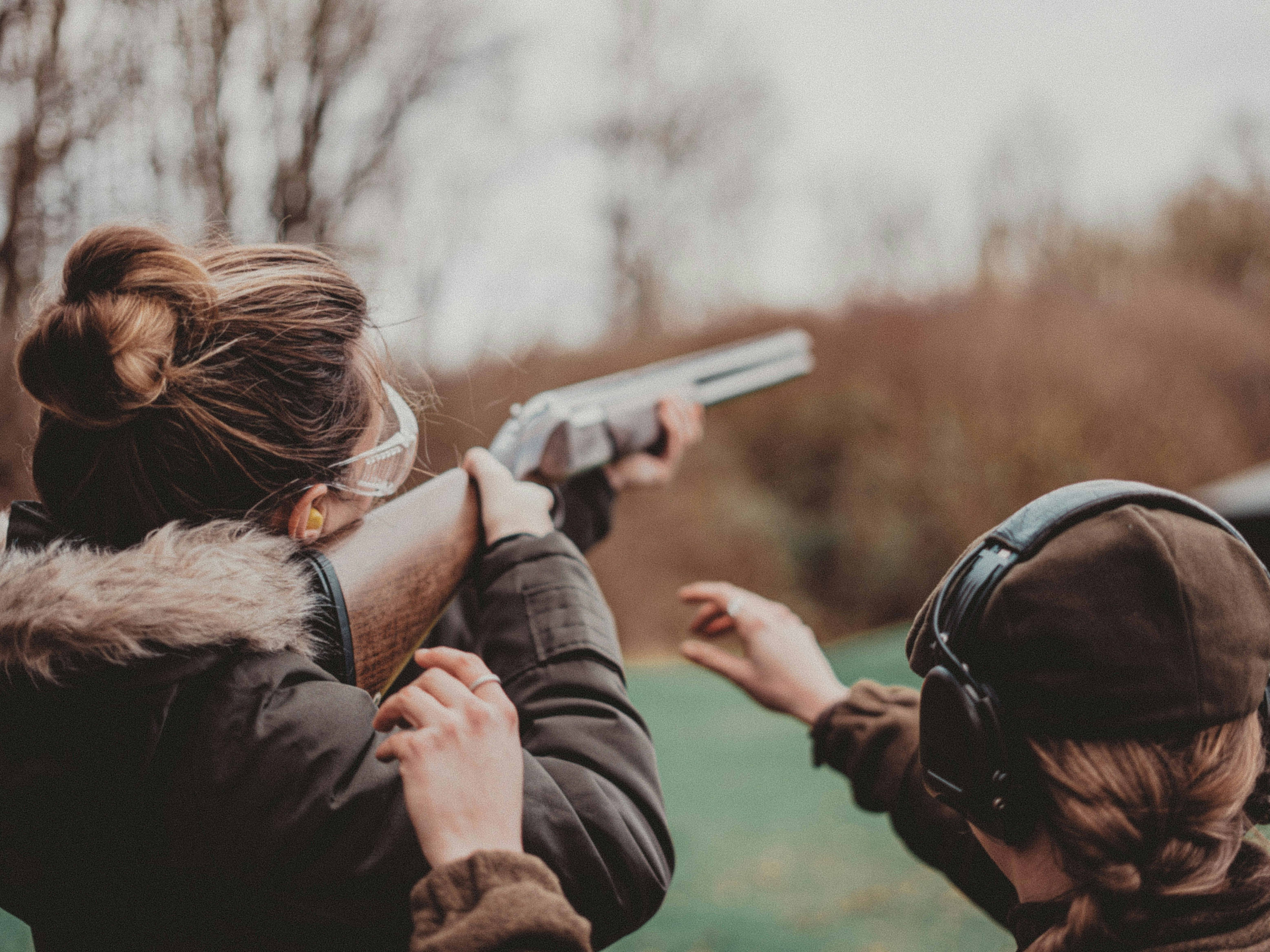 Two people outdoors, one aiming a shotgun and the other providing guidance, both wearing protective gear