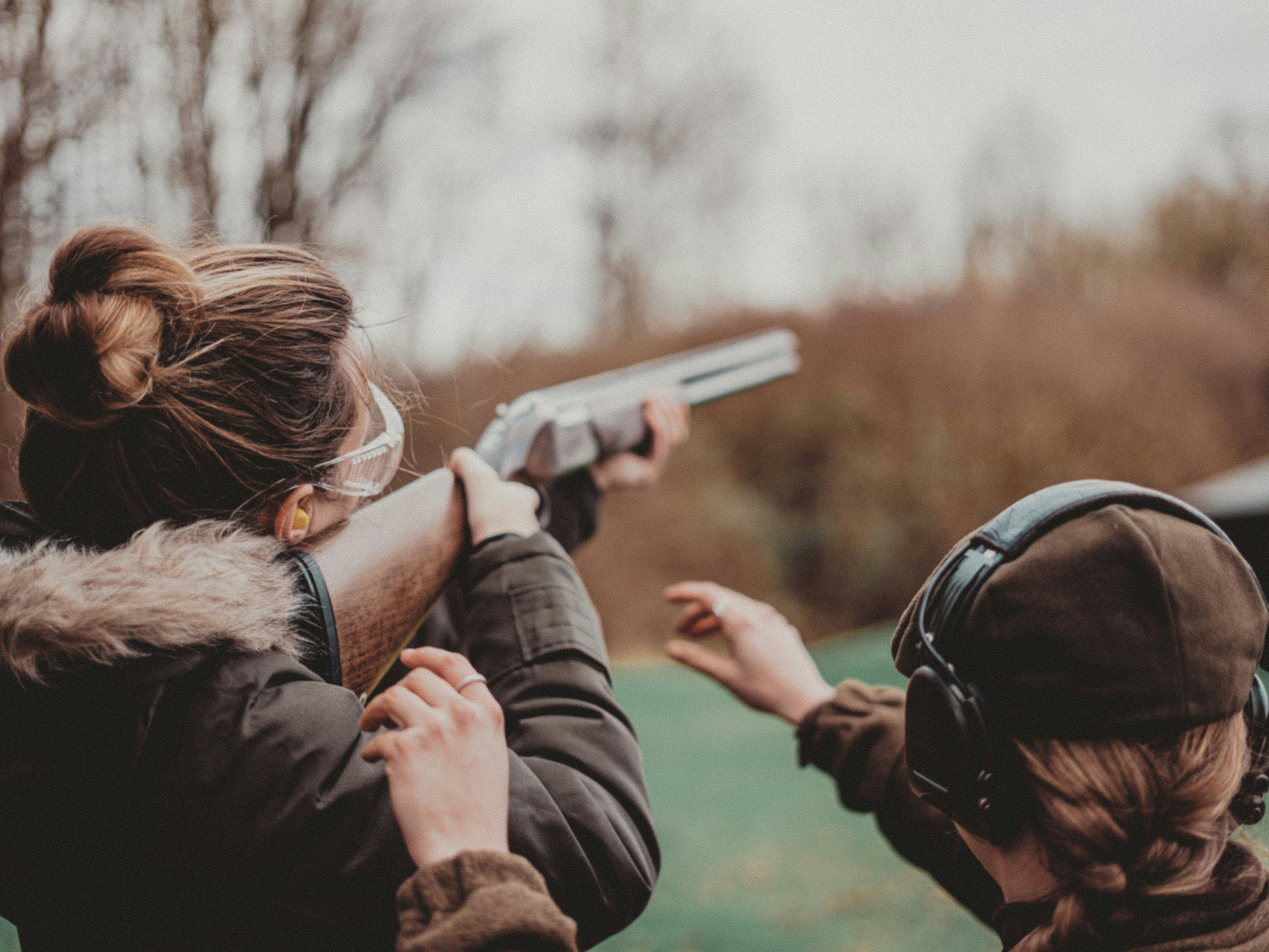 Two people outdoors, one aiming a shotgun and the other providing guidance, both wearing protective gear