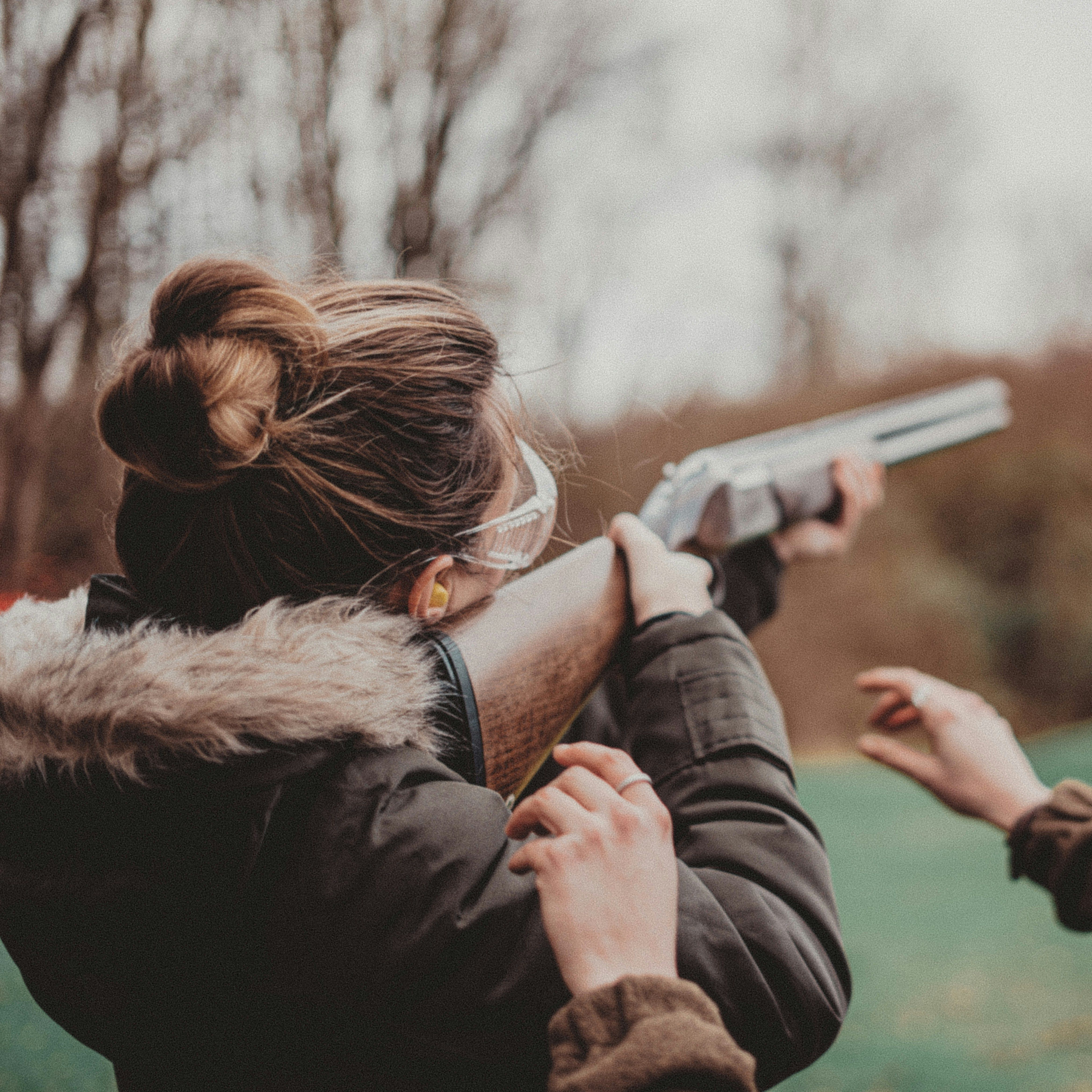 Two people outdoors, one aiming a shotgun and the other providing guidance, both wearing protective gear