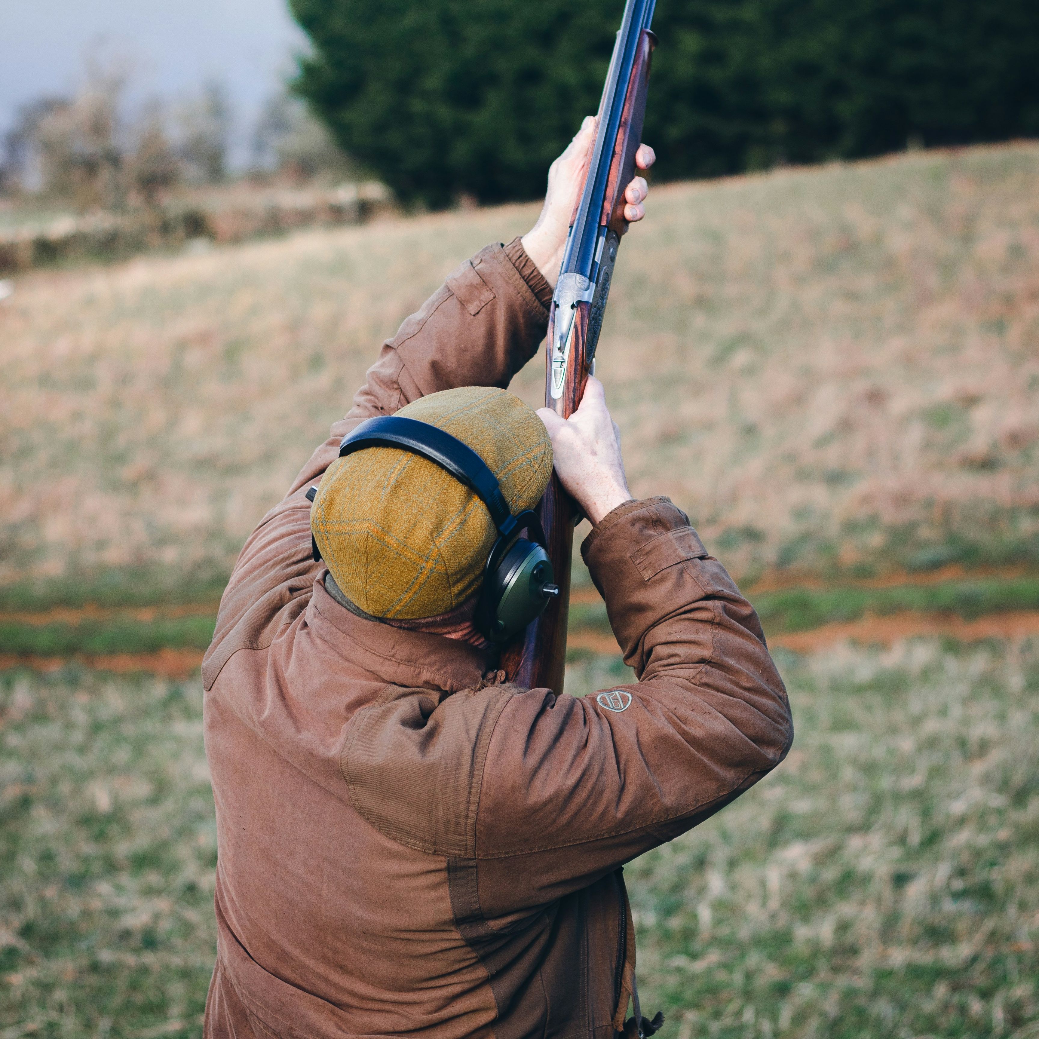 Person outdoors aiming a shotgun upwards, wearing a brown coat, ear protection, and a yellow cap.