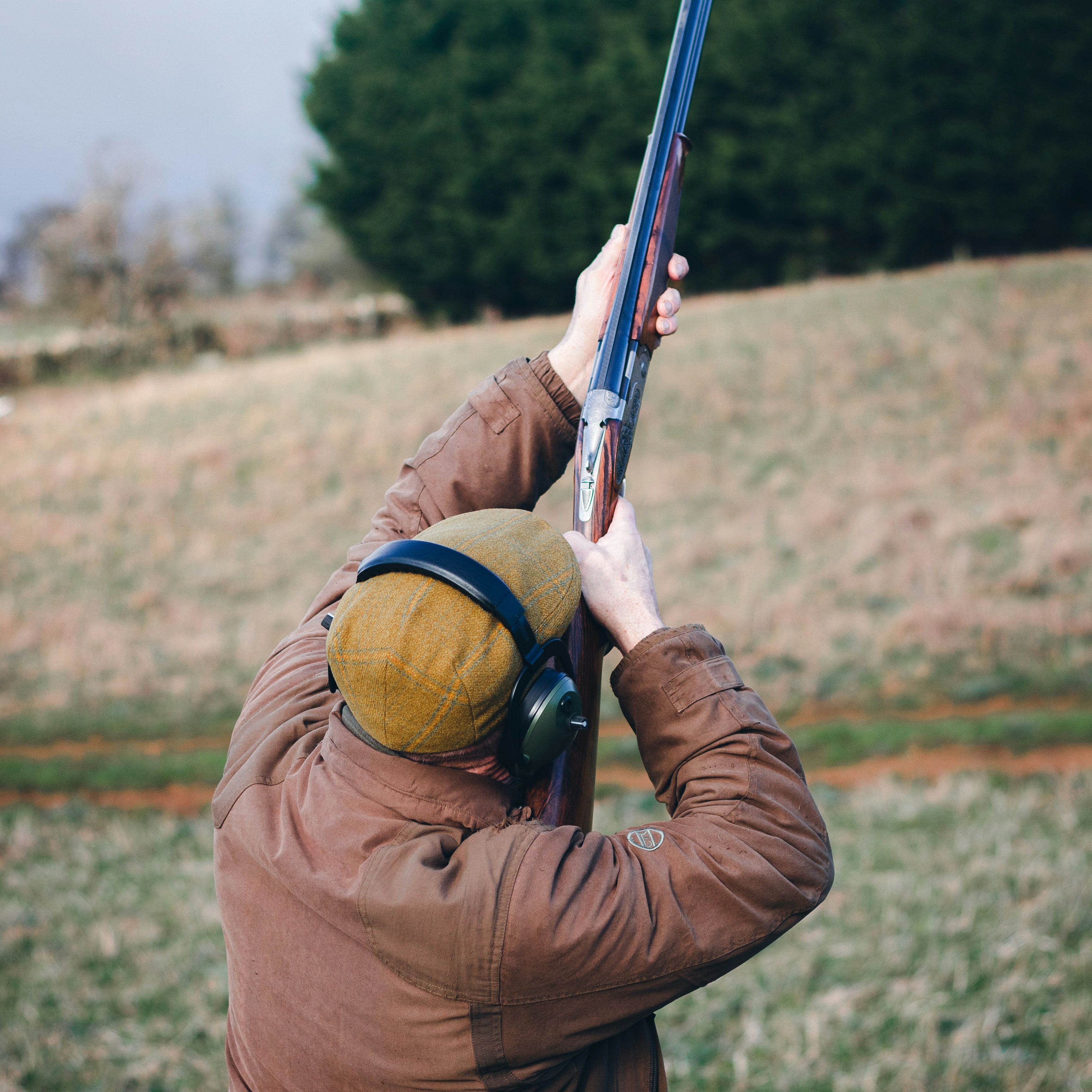 Person outdoors aiming a shotgun upwards, wearing a brown coat, ear protection, and a yellow cap.