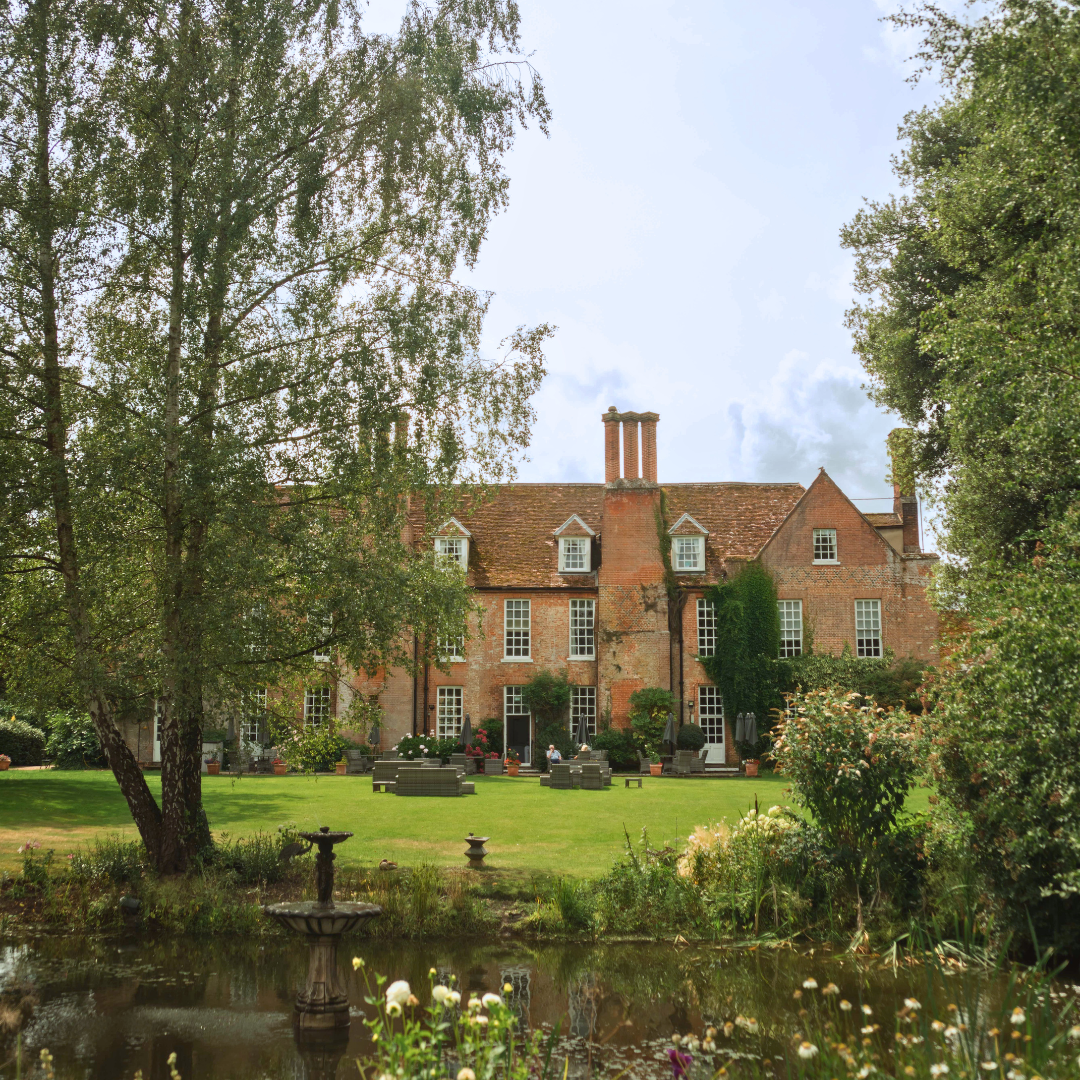 Large red-brick country house with a manicured lawn, trees and a pond in the foreground