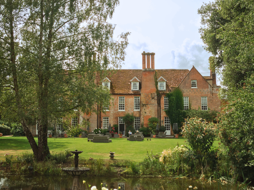 Large red-brick country house with a manicured lawn, trees and a pond in the foreground