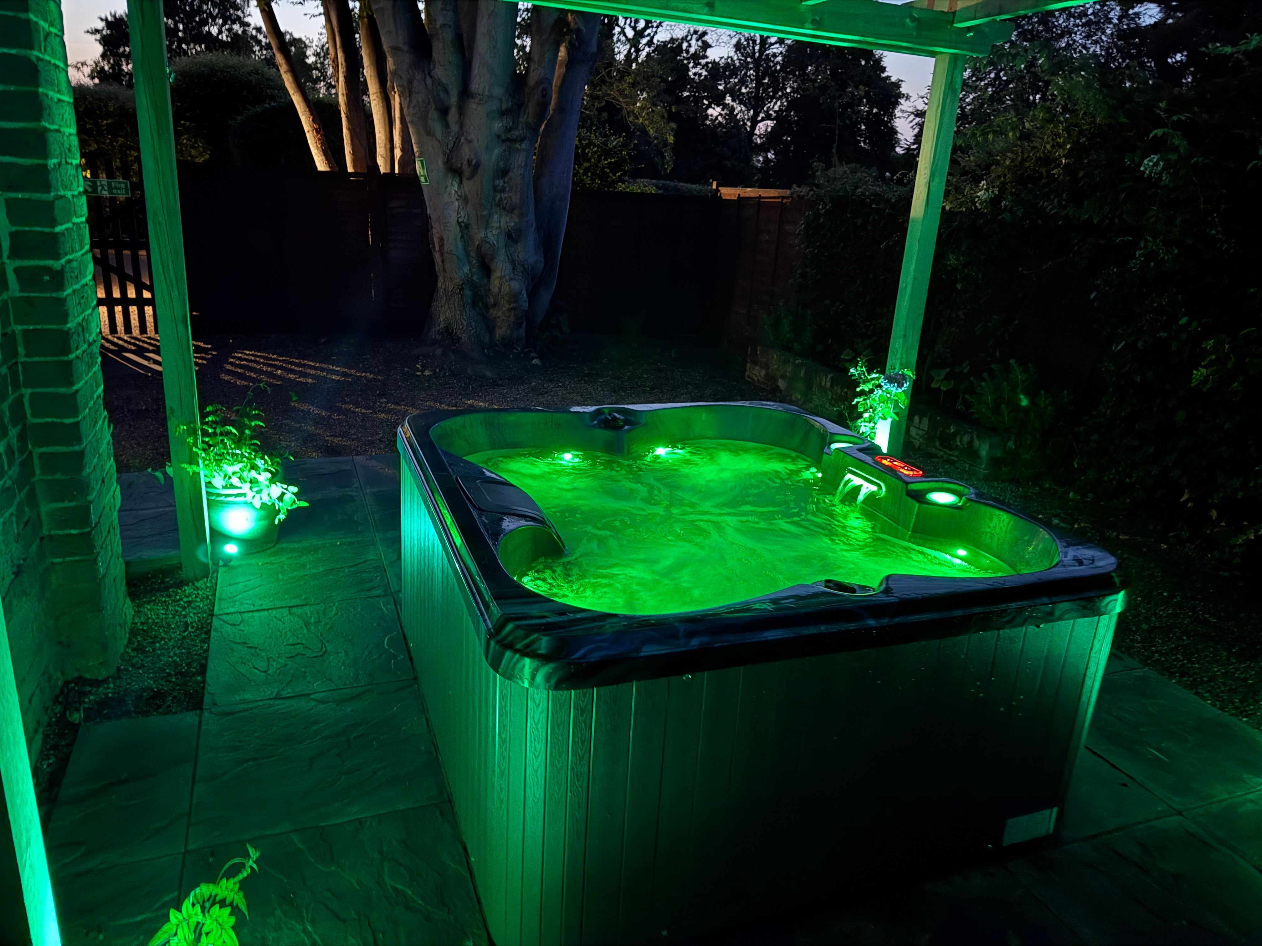 Outdoor hot tub glowing green under a pergola at dusk, surrounded by plants and trees