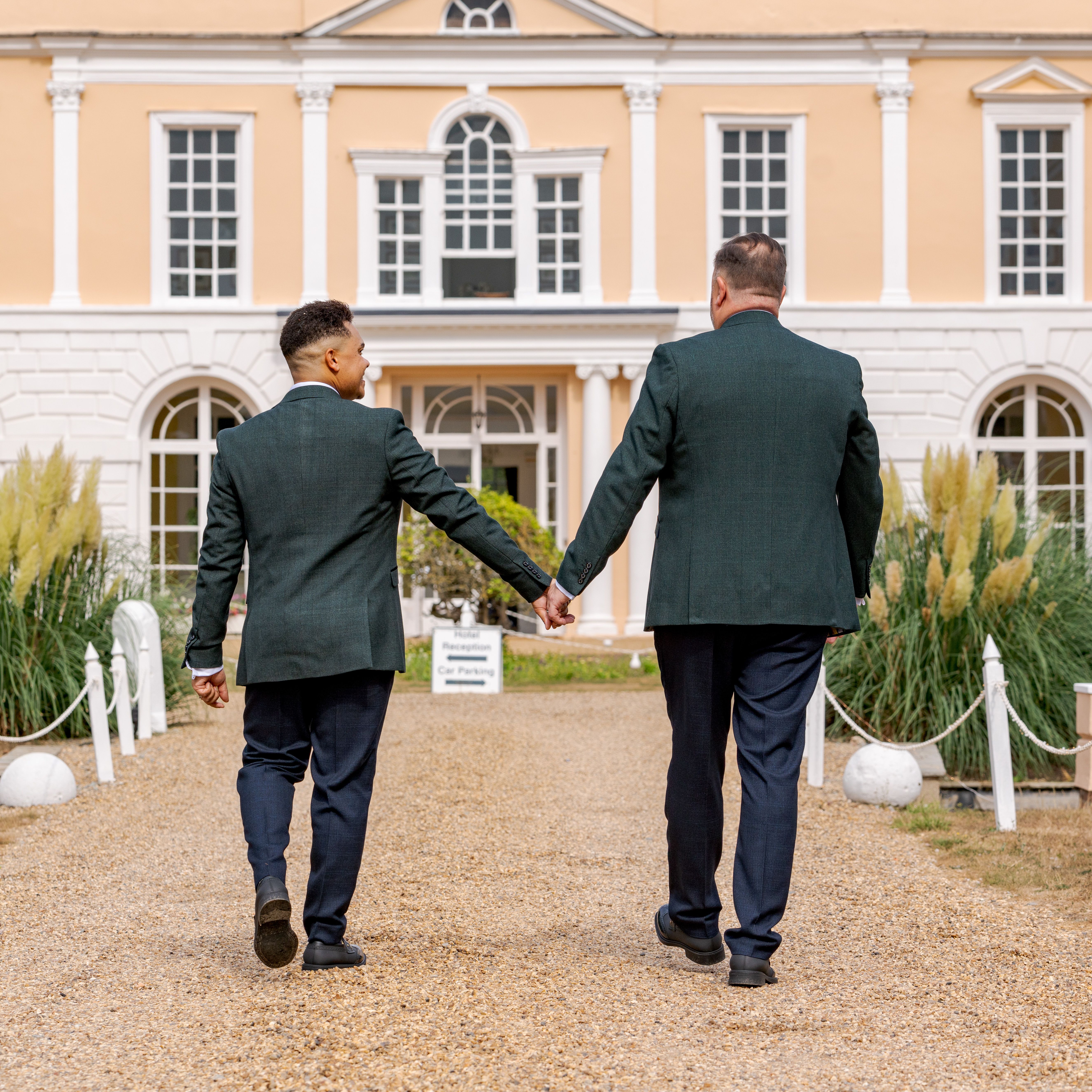 Two grooms holding hands walking towards a grand country house wedding venue in daylight