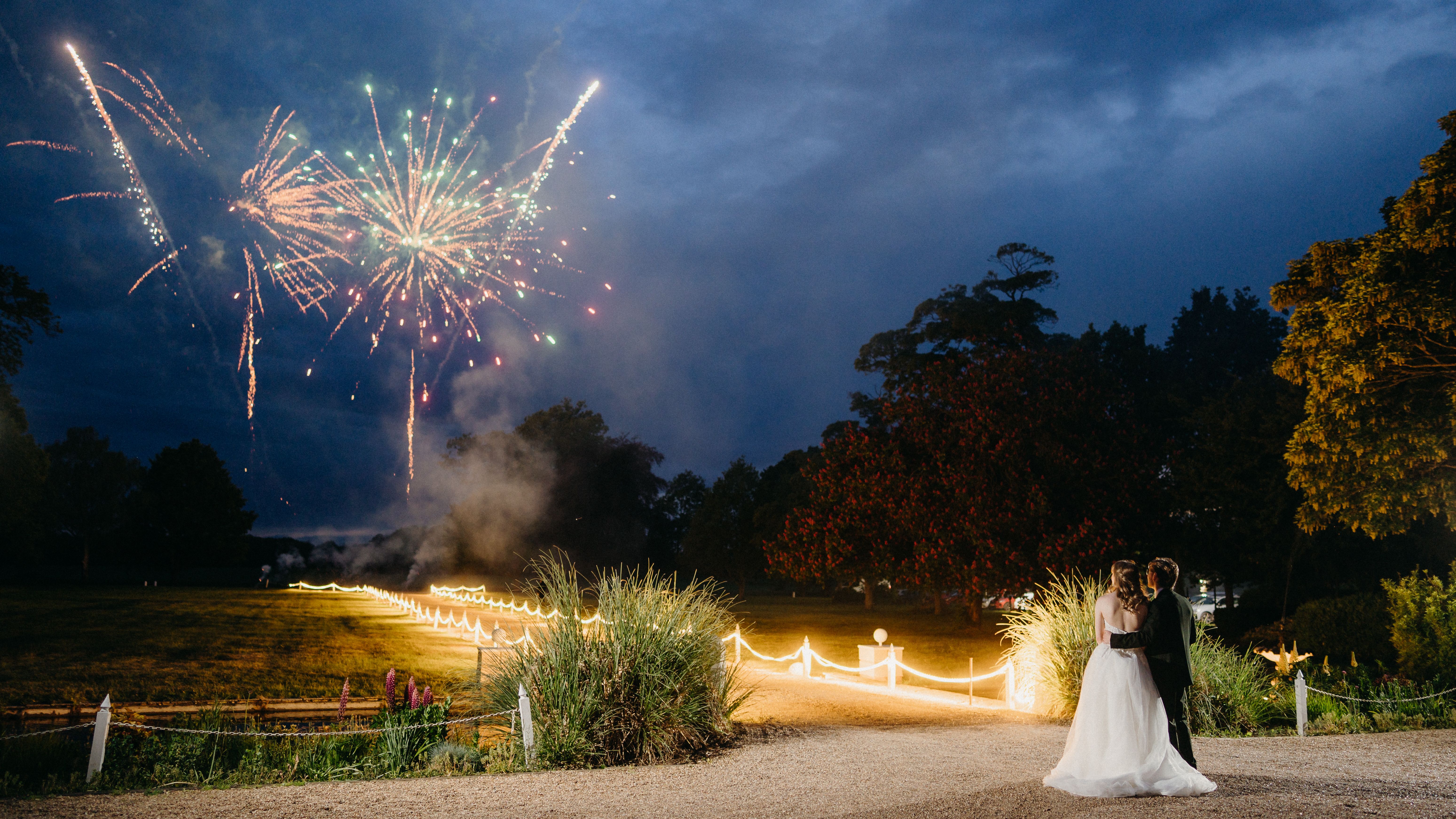 Bride and groom embracing on a lit garden path as fireworks burst in the night sky