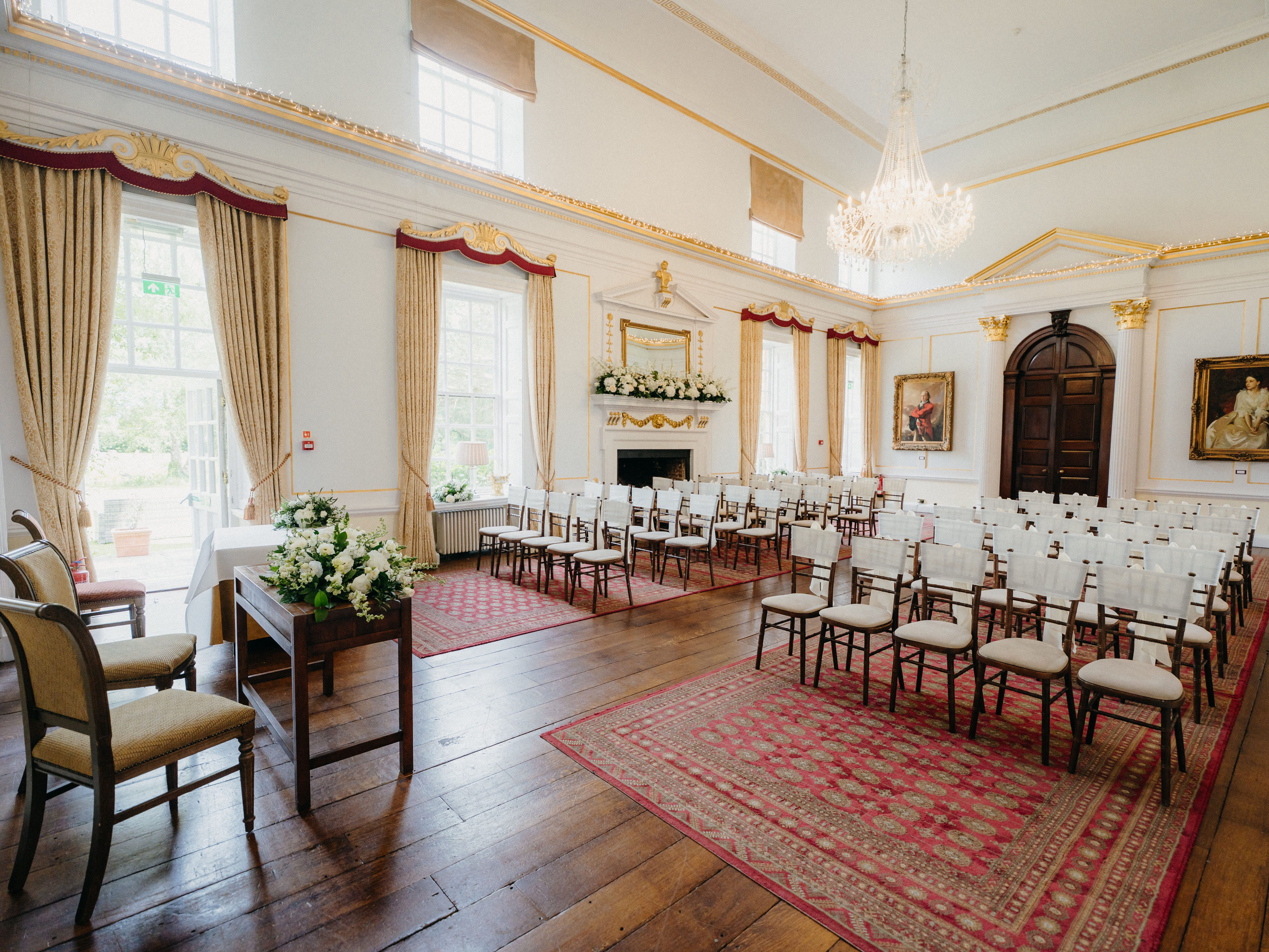 Elegant wedding ceremony room with chandelier, tall windows and rows of white chairs on red patterned rugs