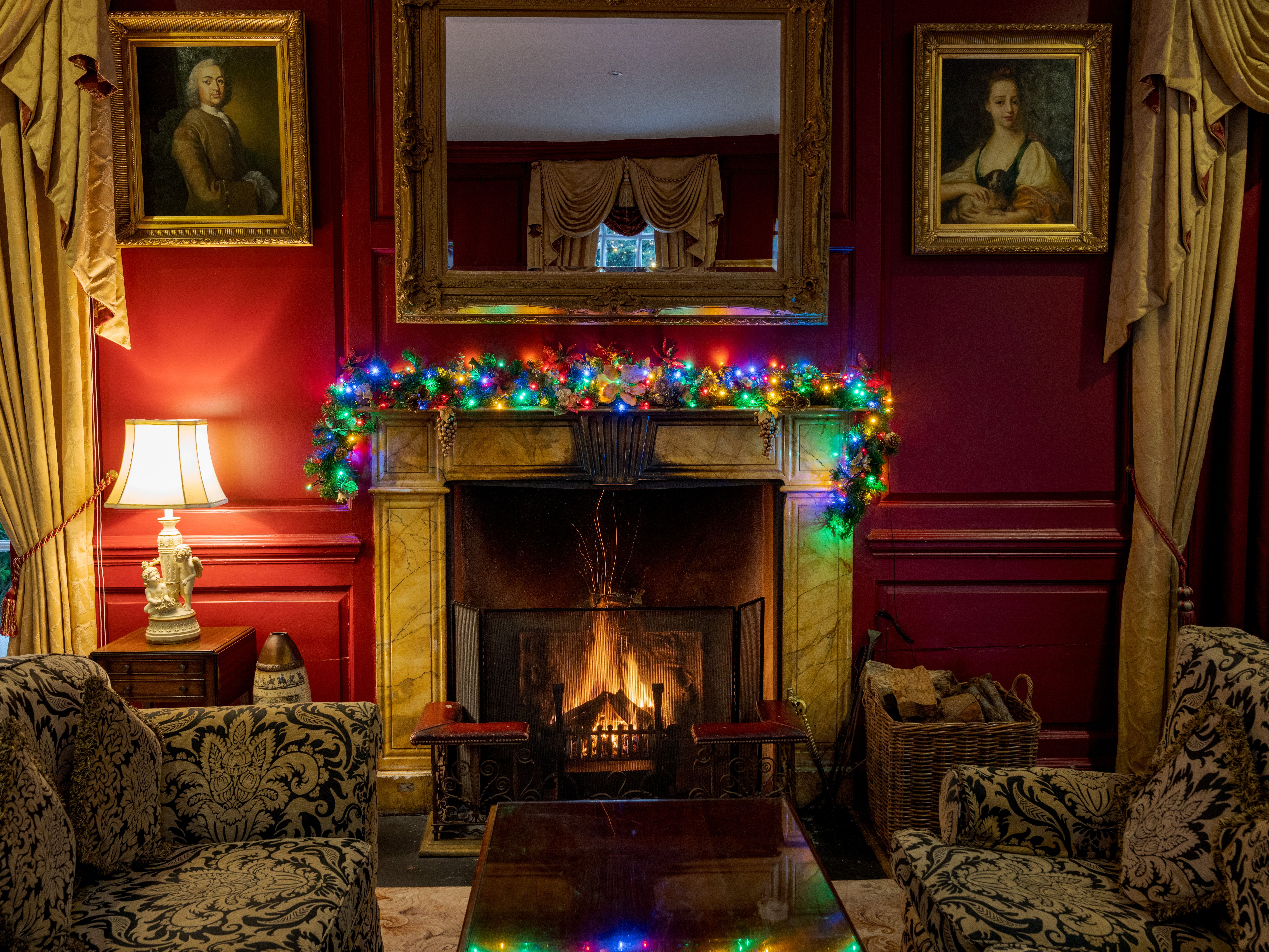 Cosy sitting room with a lit fireplace, red walls, gilt-framed portraits and Christmas garland with coloured lights on the mantelpiece