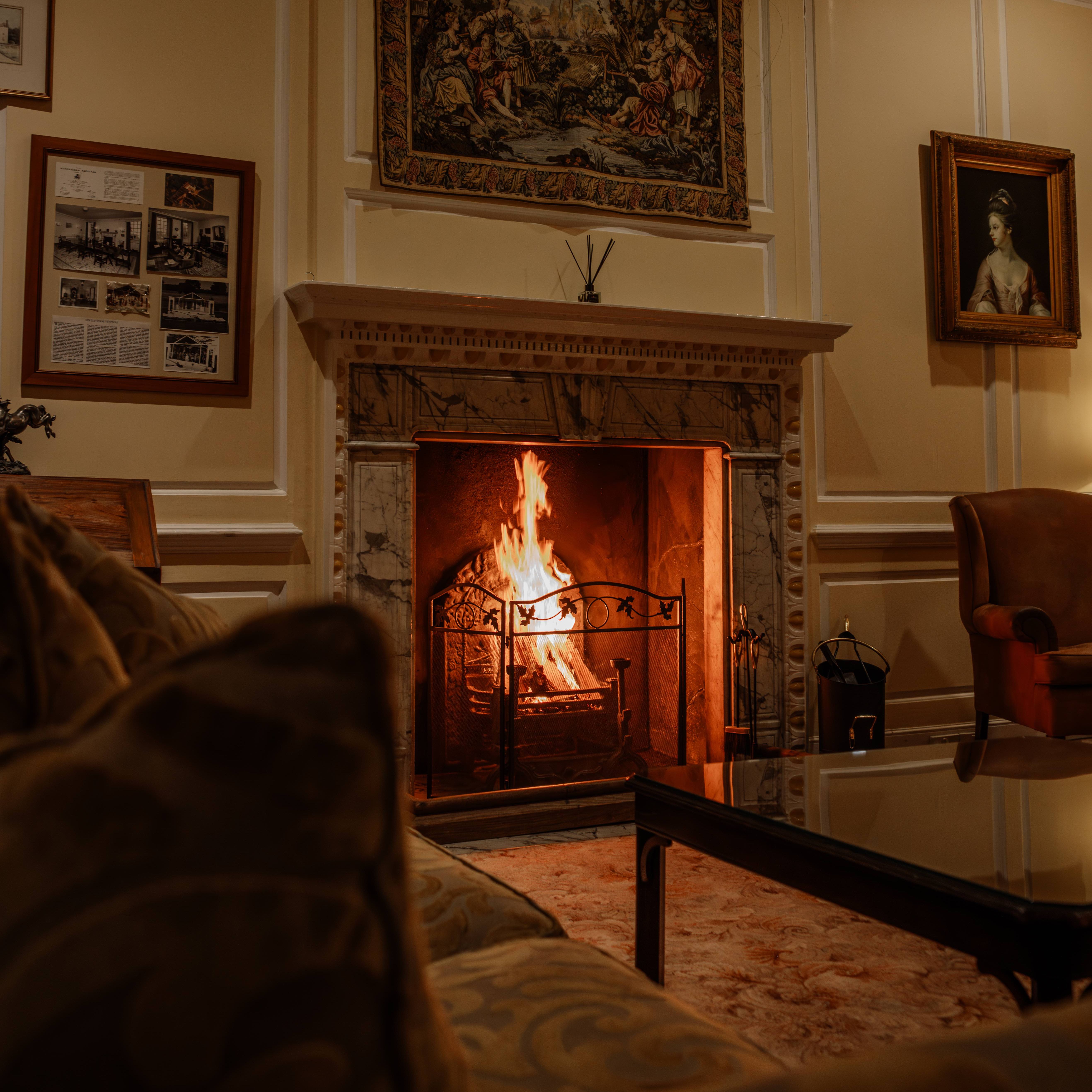 Cozy living room with a lit fireplace, ornate mantle, classic paintings, and comfortable seating.