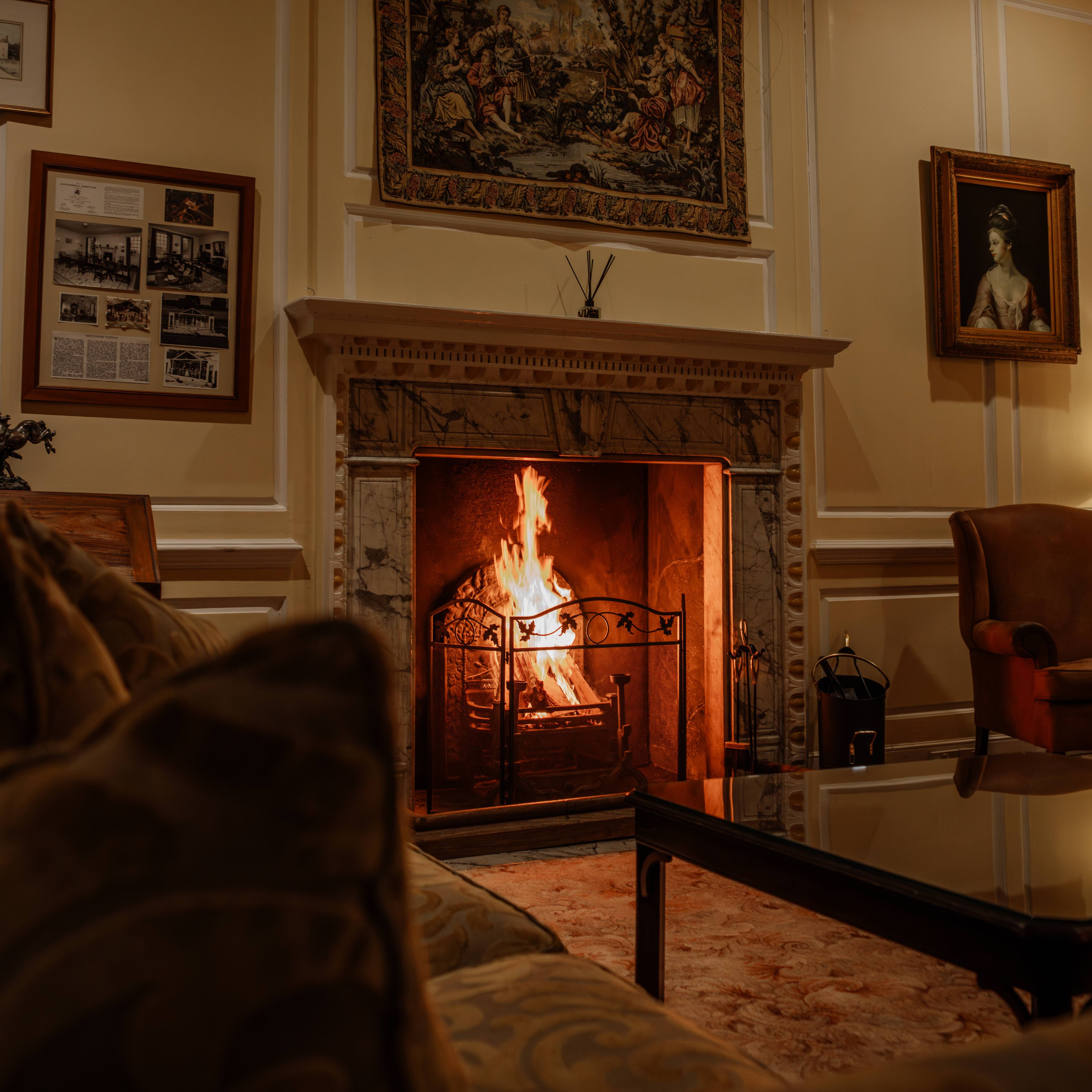 Cozy living room with a lit fireplace, ornate mantle, classic paintings, and comfortable seating.