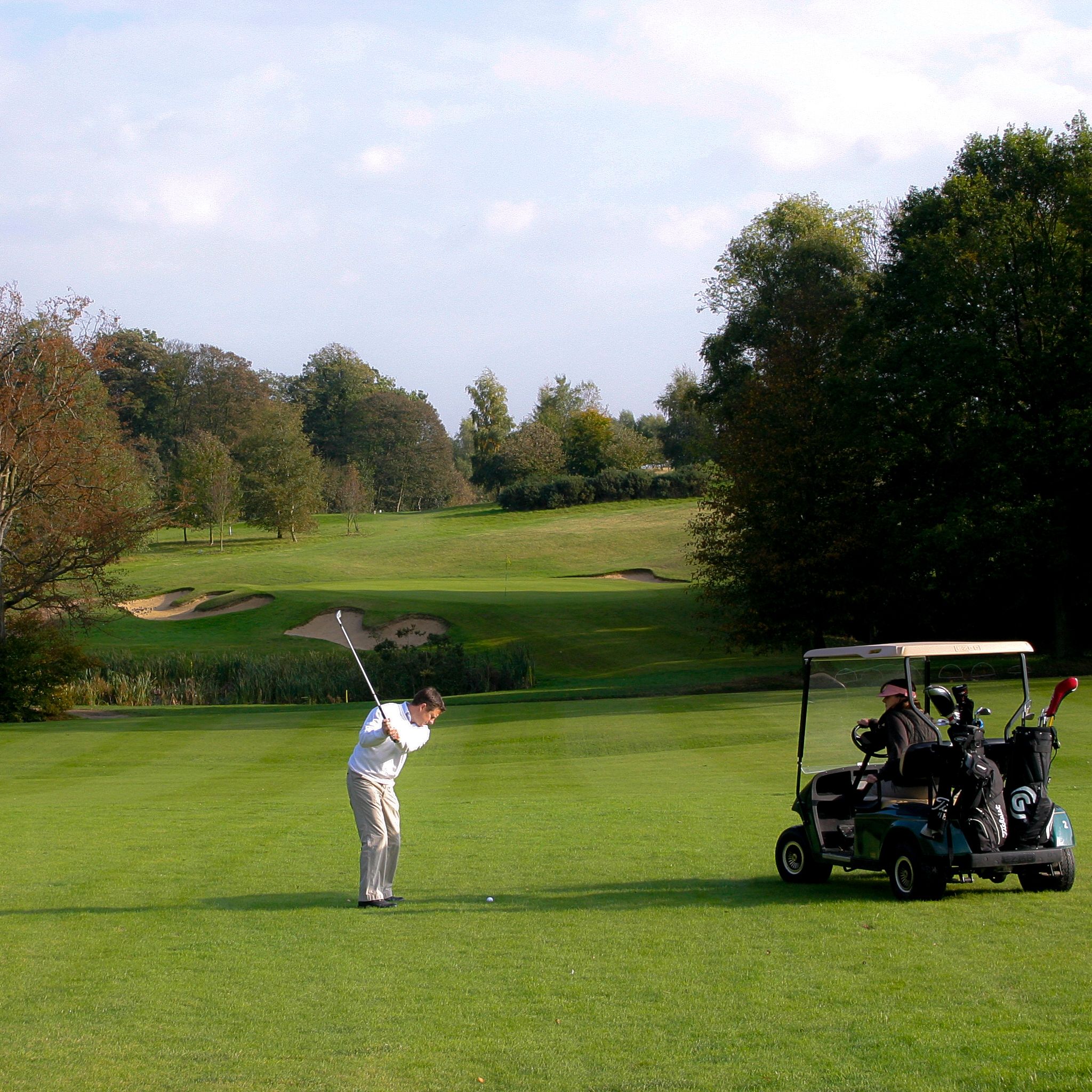 Golfer swinging on a tree-lined fairway beside a golf buggy