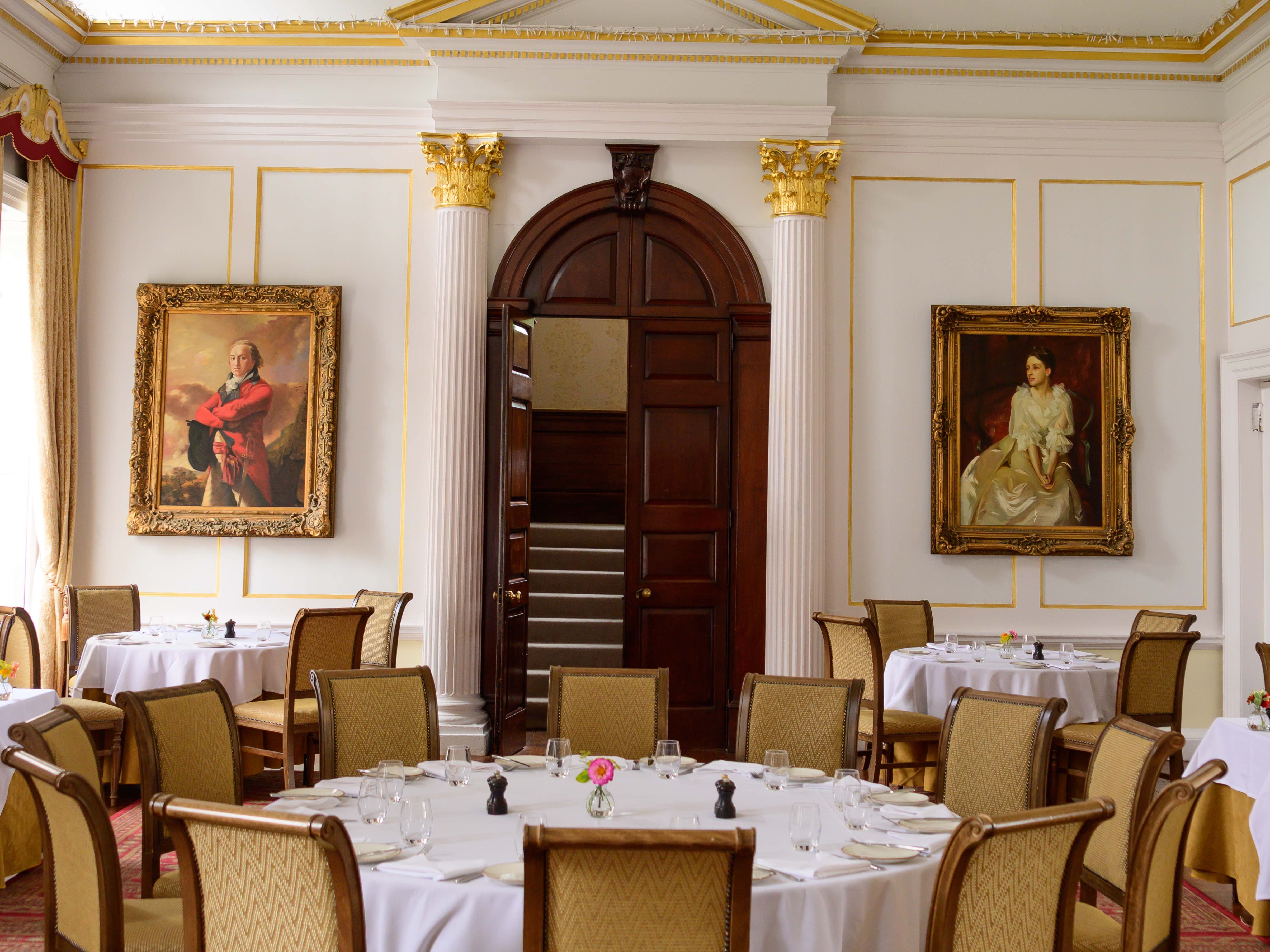 Elegant dining room with classic decor, round tables, a chandelier, and portrait paintings on the walls
