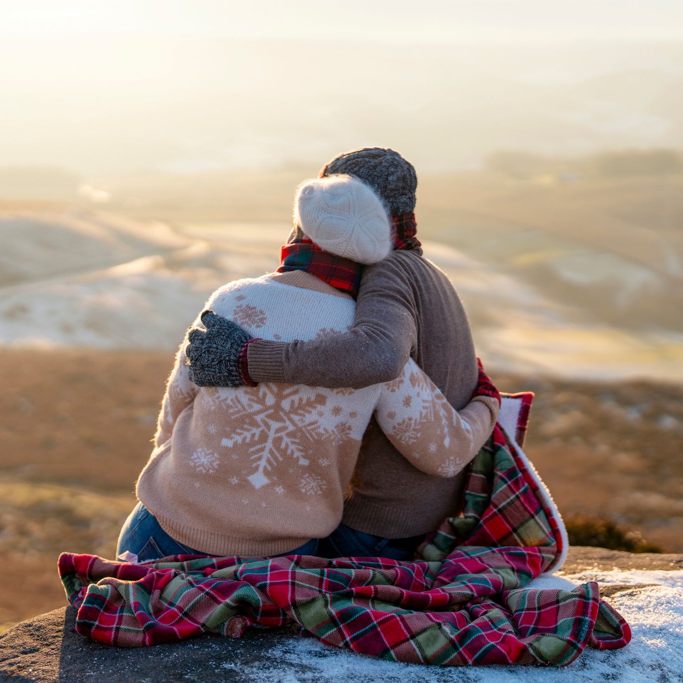 A couple dressed in warm winter clothing sits on a stone ledge, wrapped in a red and green plaid blanket, embracing each other while gazing at a snow-dusted valley at sunset.