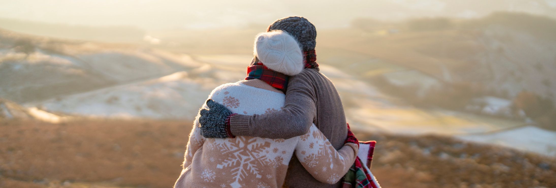 A couple dressed in warm winter clothing sits on a stone ledge, wrapped in a red and green plaid blanket, embracing each other while gazing at a snow-dusted valley at sunset.